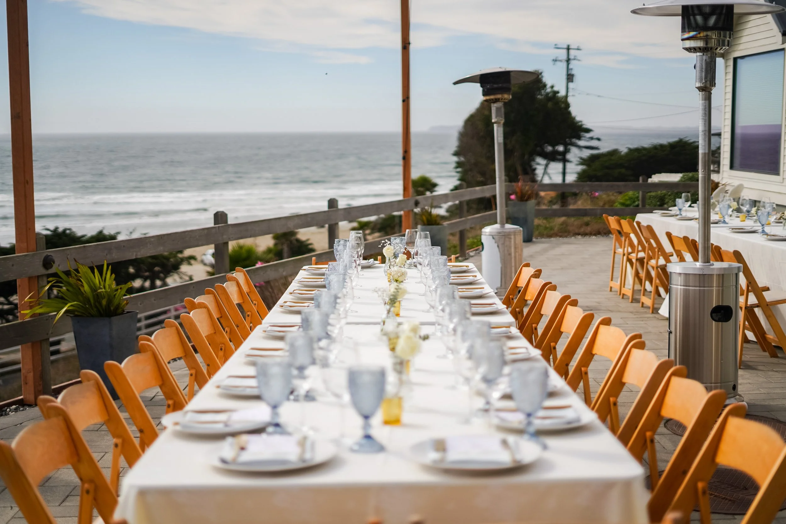 A long outdoor dining table set with white tablecloth, arranged with glassware, white plates, and floral centerpieces, on a deck with a view of the ocean and a cloudy sky, flanked by orange chairs and patio heaters.