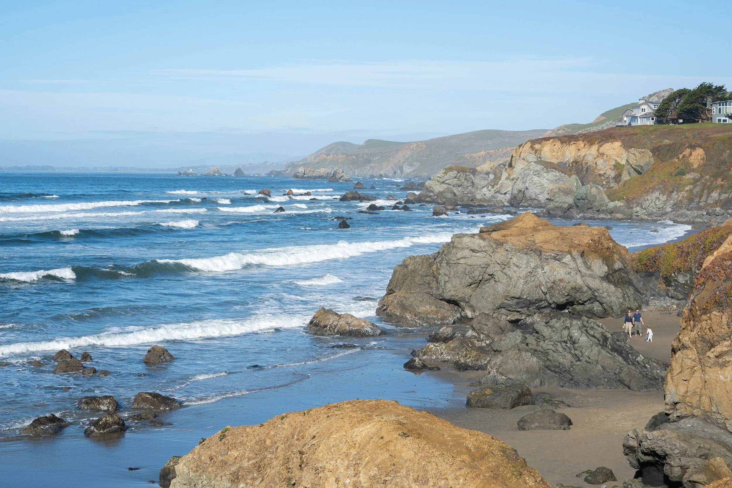 View of a coastal area with rocky cliffs, waves crashing onto the shore, and houses on top of the cliffs. A few people are walking along the beach.