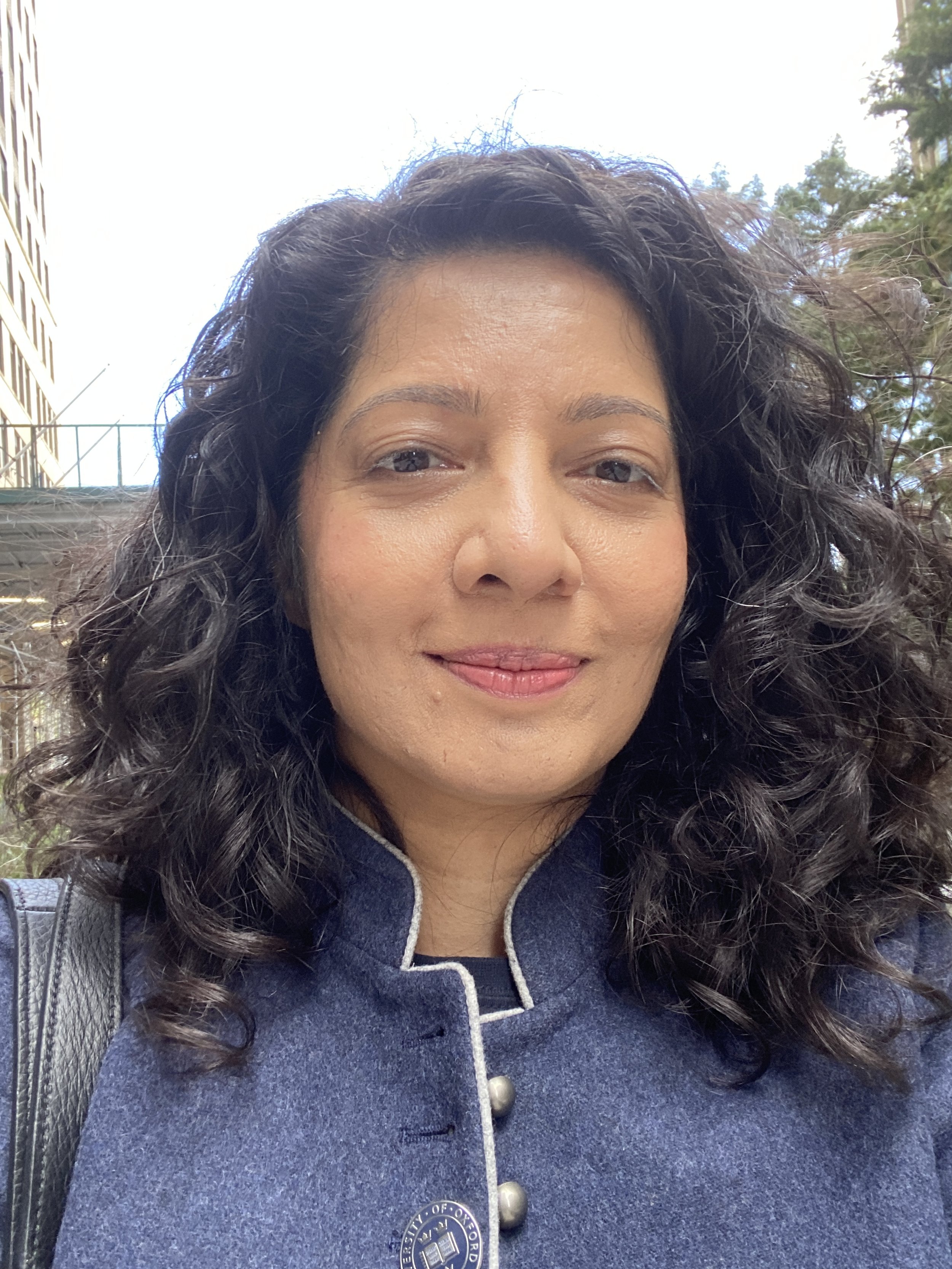 Close-up of a woman with curly dark hair, wearing a blue coat with silver buttons, outdoors on a cloudy day.