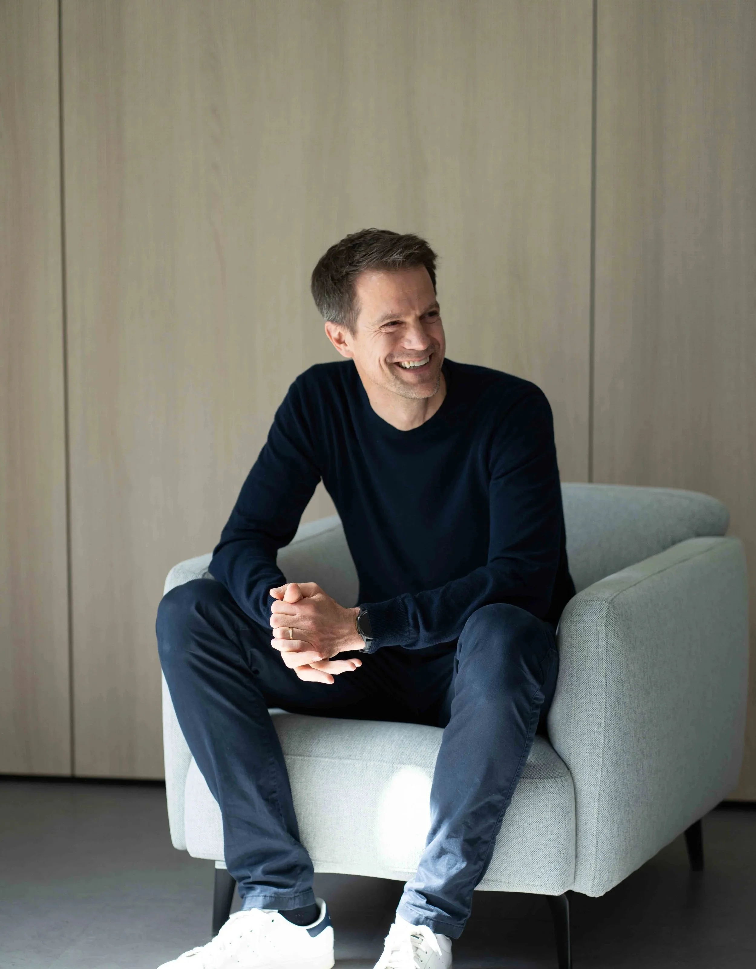 Ein lachender Mann sitzt auf einem grauen Sofa in einem Raum mit hellen Wänden.