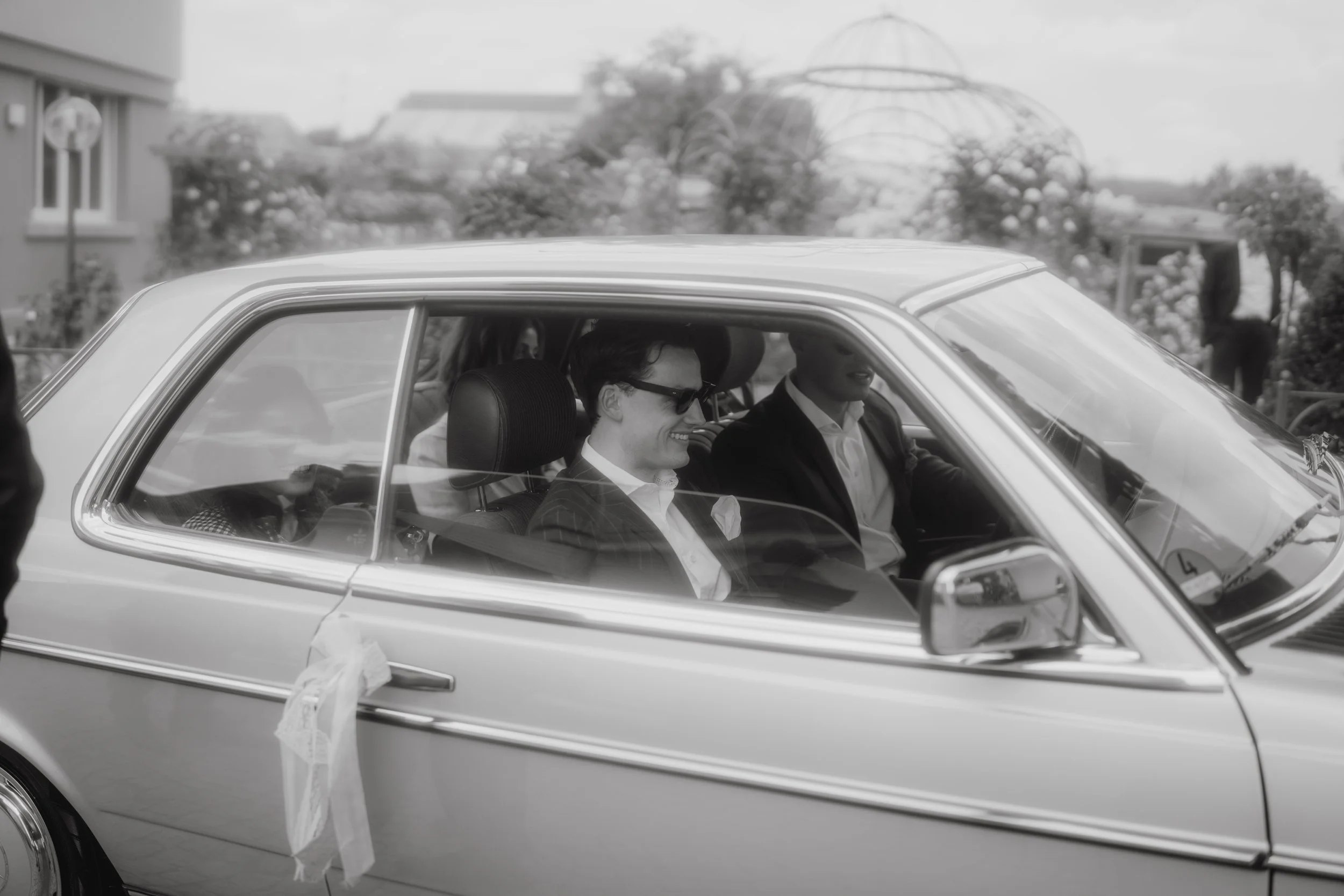 Zwei Männer sitzen in einem klassischen weißen Auto, vermutlich bei einer besonderen Gelegenheit wie einer Hochzeit, während die Fenster des Autos mit Bändern dekoriert sind.