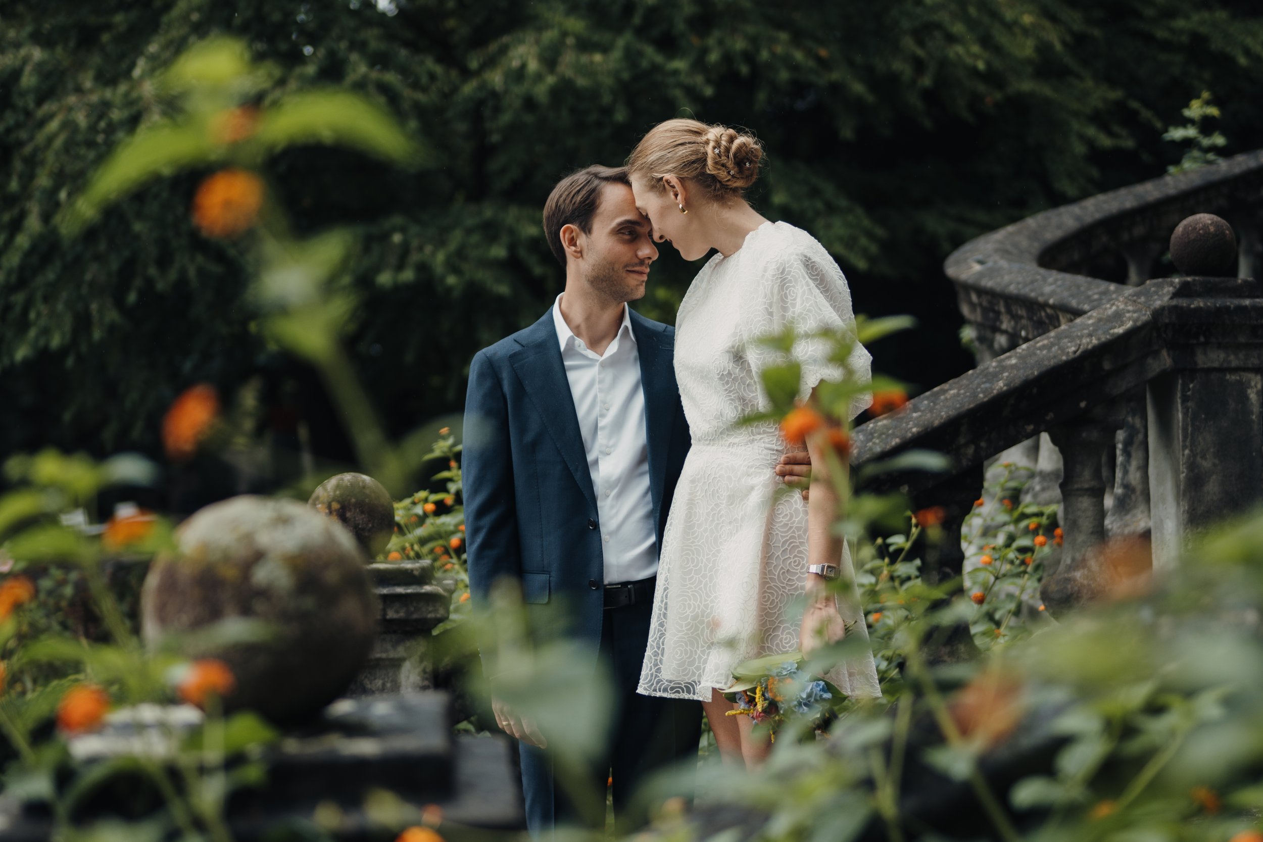 Ein Paar, der Mann trägt einen blauen Anzug, die Frau trägt ein weißes Kleid, stehen sich mit Stirn an Stirn auf einer Gartenbalkon mit Ranken und Blumen im Hintergrund.