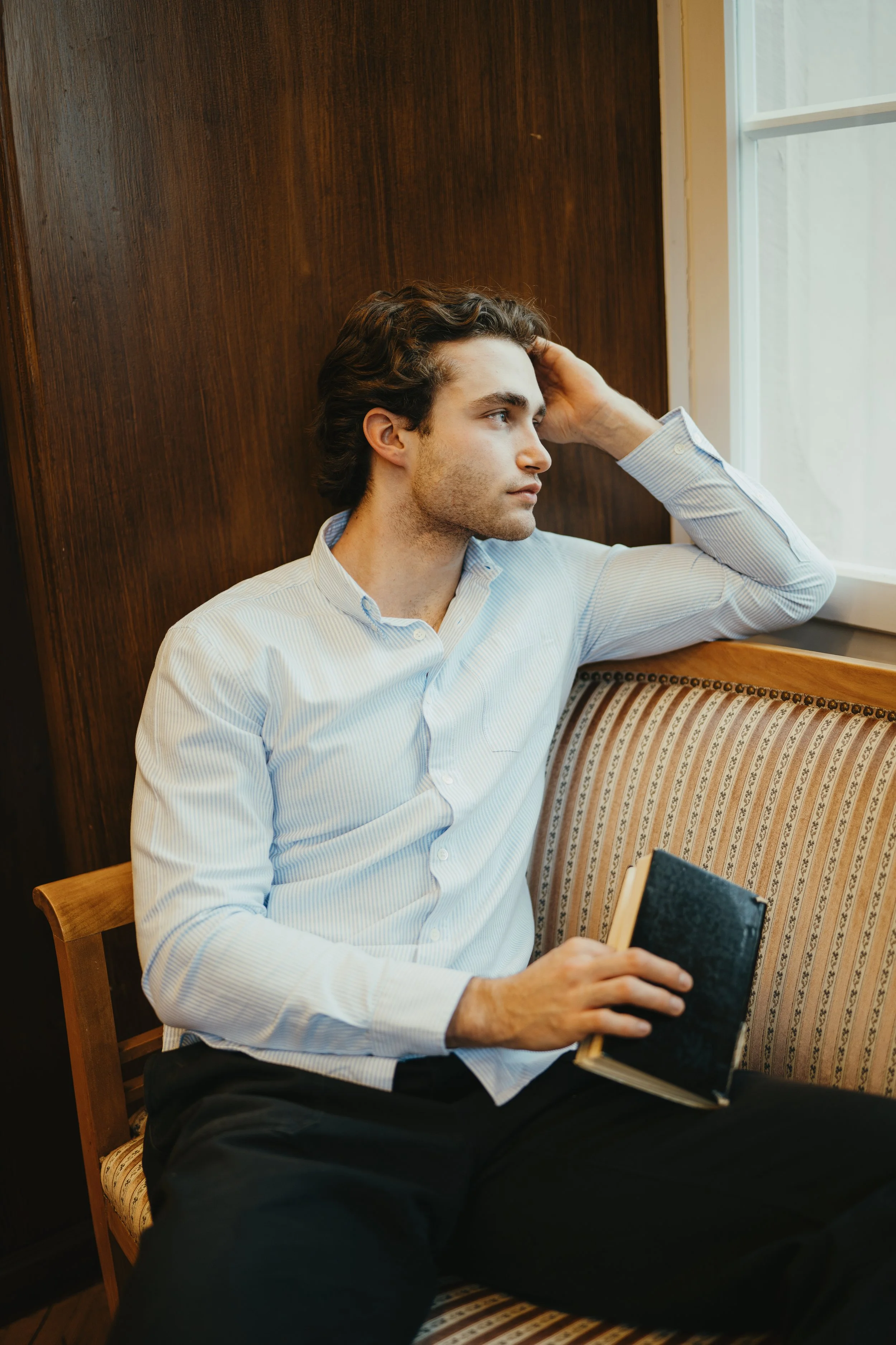 Junger Mann sitzt in einem Raum neben einem Fenster, hält ein Buch in der Hand und sieht nachdenklich aus.