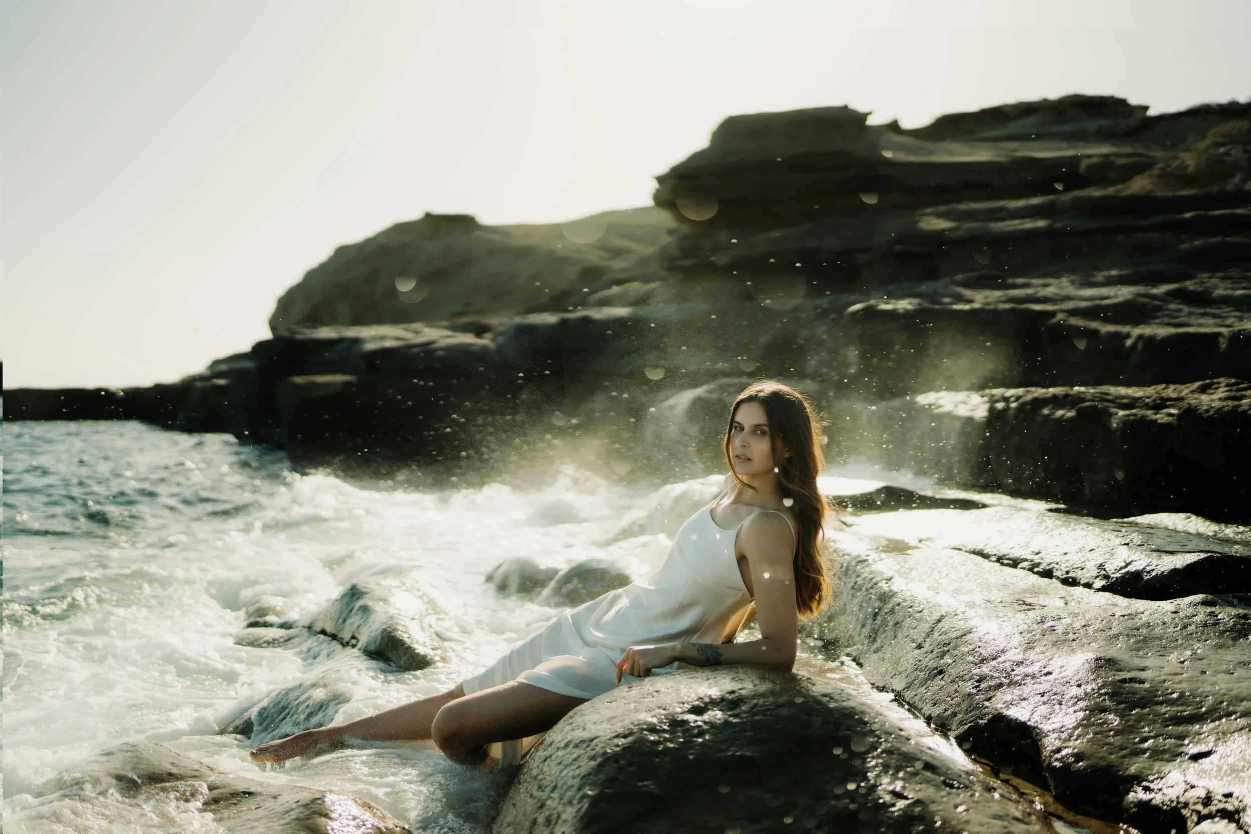 Eine Frau in einem weißen Kleid liegt am Strand auf den Felsen, mit Wasser umgeben und Klippen im Hintergrund, bei Sonnenuntergang oder Sonnenaufgang.