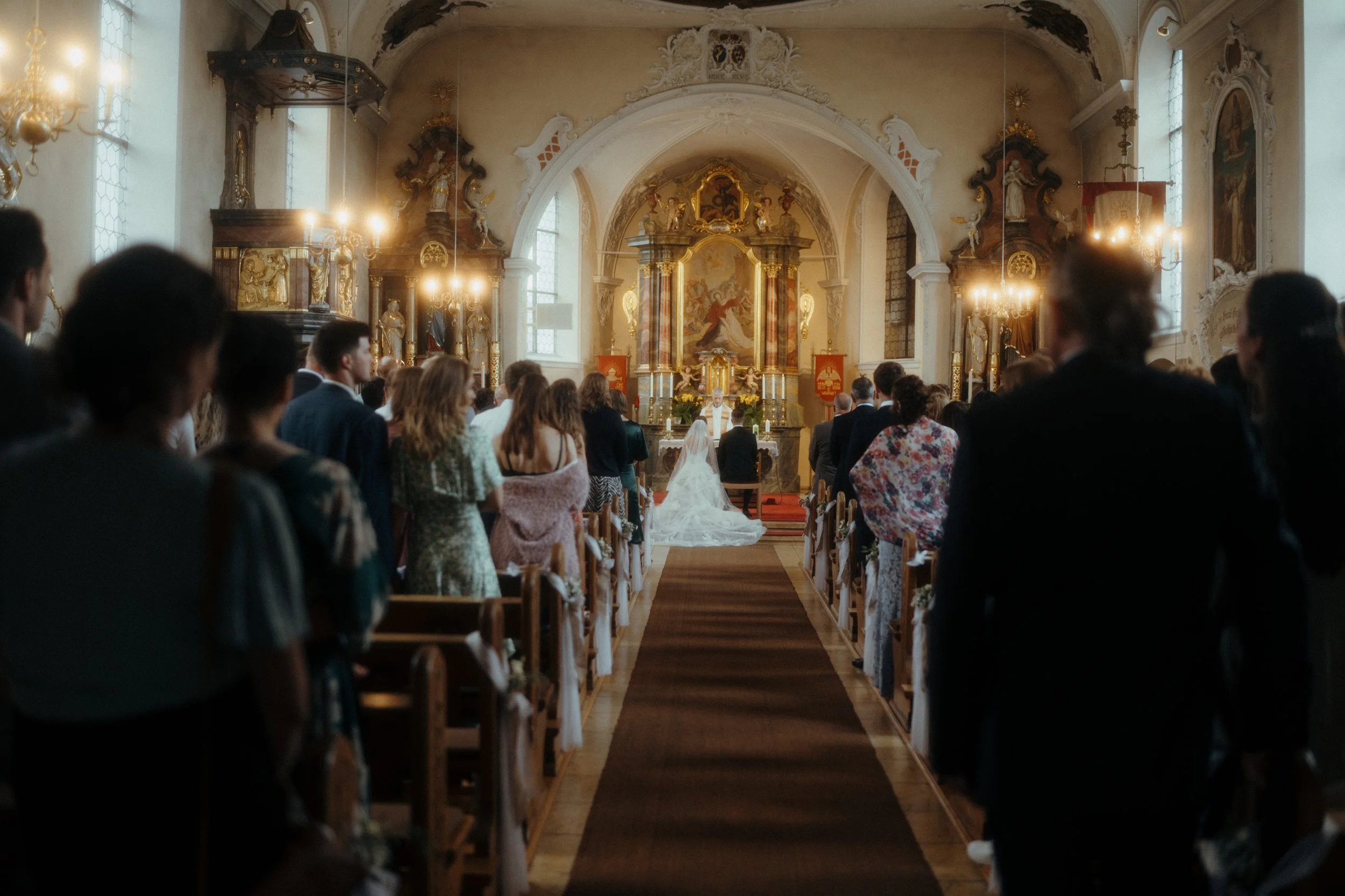 Eine Gruppe von Menschen, die in einer Kirche bei einer Hochzeit oder einem Gottesdienst sitzen, mit Blick auf den Altar im Hintergrund.