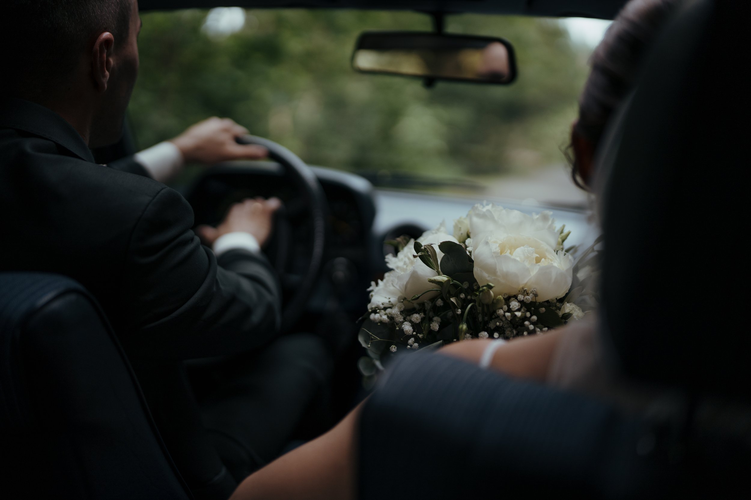 Ein Mann fährt in einem Auto, während eine Frau daneben sitzt. Auf dem Rücksitz liegt ein Brautstrauß aus weißen Blumen.