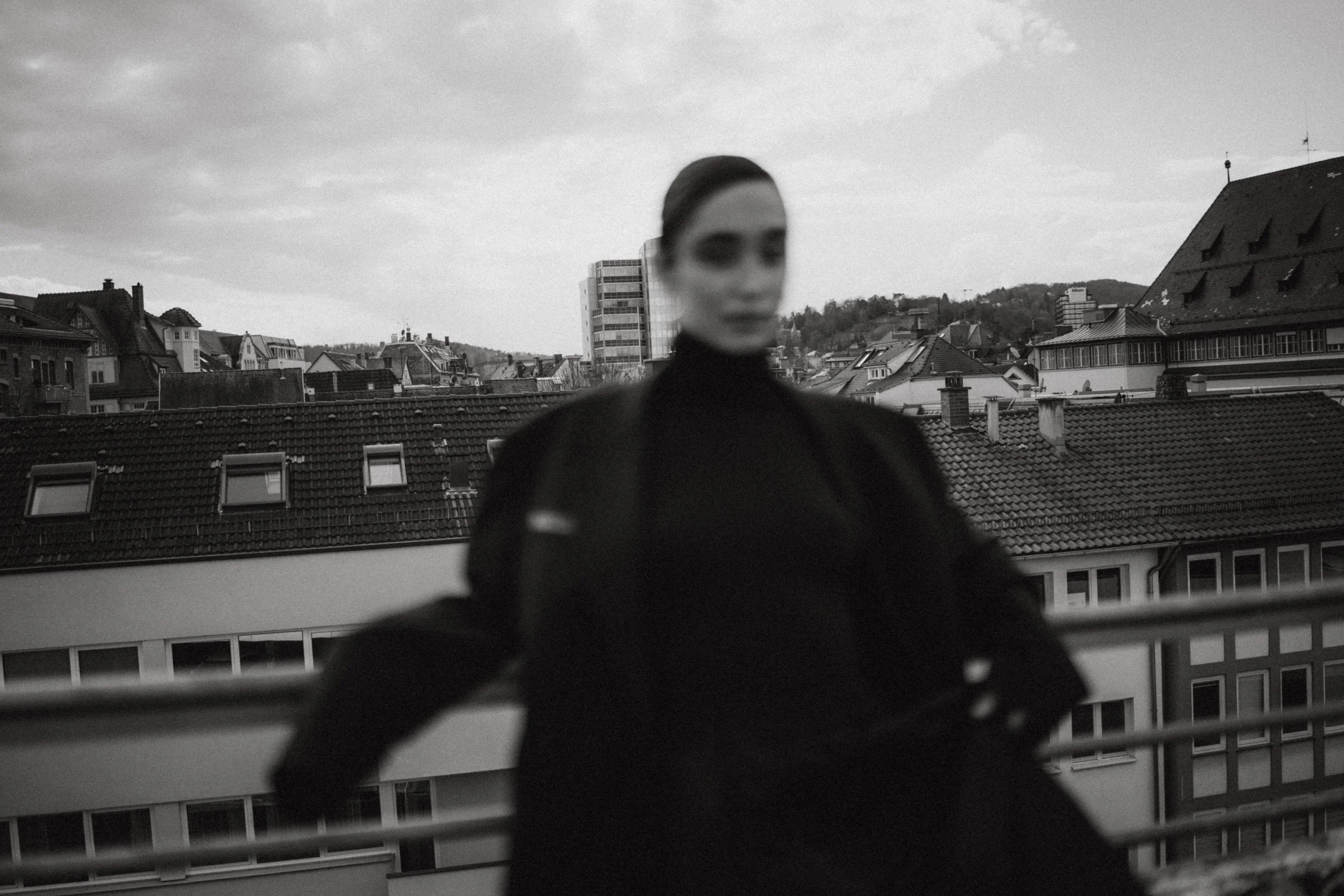 Schwarzer Mensch mit dunklen Haaren und Sonnenbrille steht auf einem Balkon mit Stadtblick, im Hintergrund Häuser und Hügel bei bewölktem Himmel, Schwarz-Weiß-Foto.