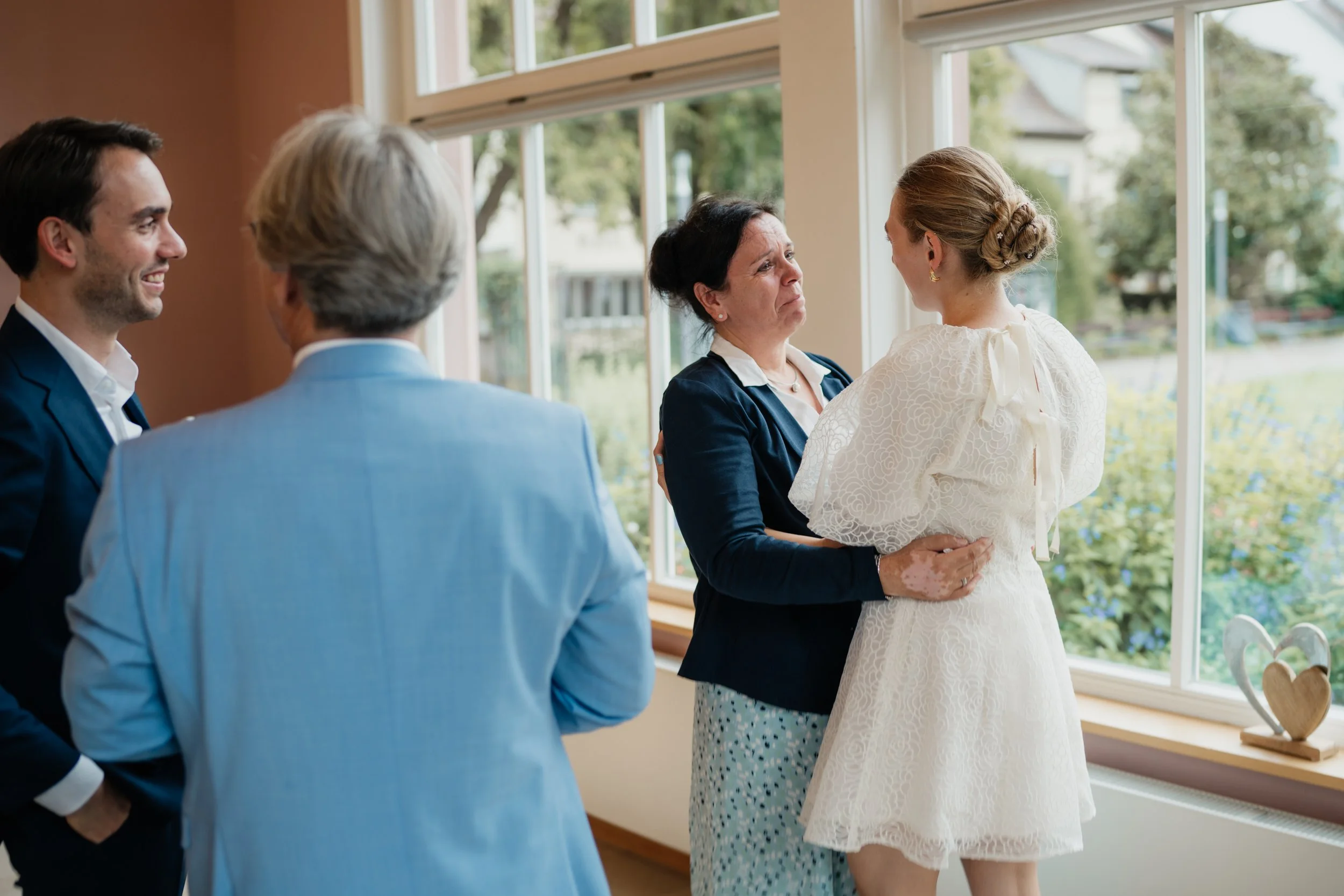 Eine Trauung im Haus, bei der eine Braut mit Schleier und Kleid von einem Mädchen mit integriertem Kleid und ihrer Mutter umarmt wird, während drei Männer zuschauen.