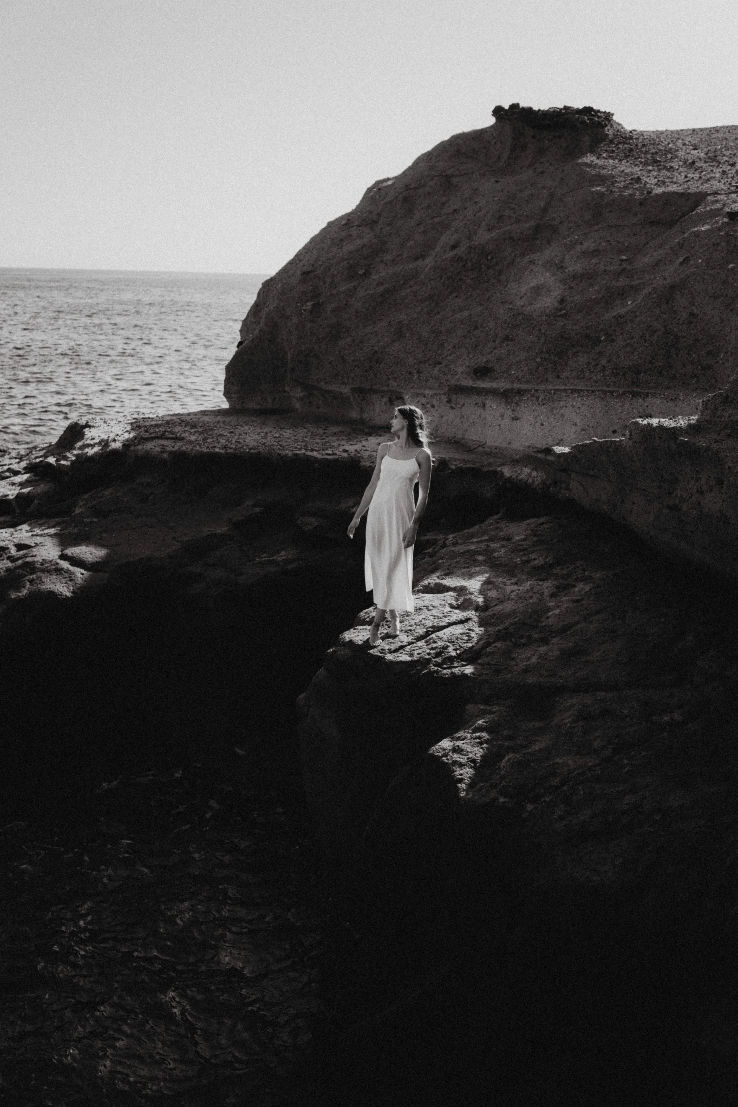 Eine Frau in einem weißen Kleid steht auf Felsen am Meer. Es ist eine Schwarz-Weiß-Aufnahme und die Szenerie wirkt ruhig und meditativ.