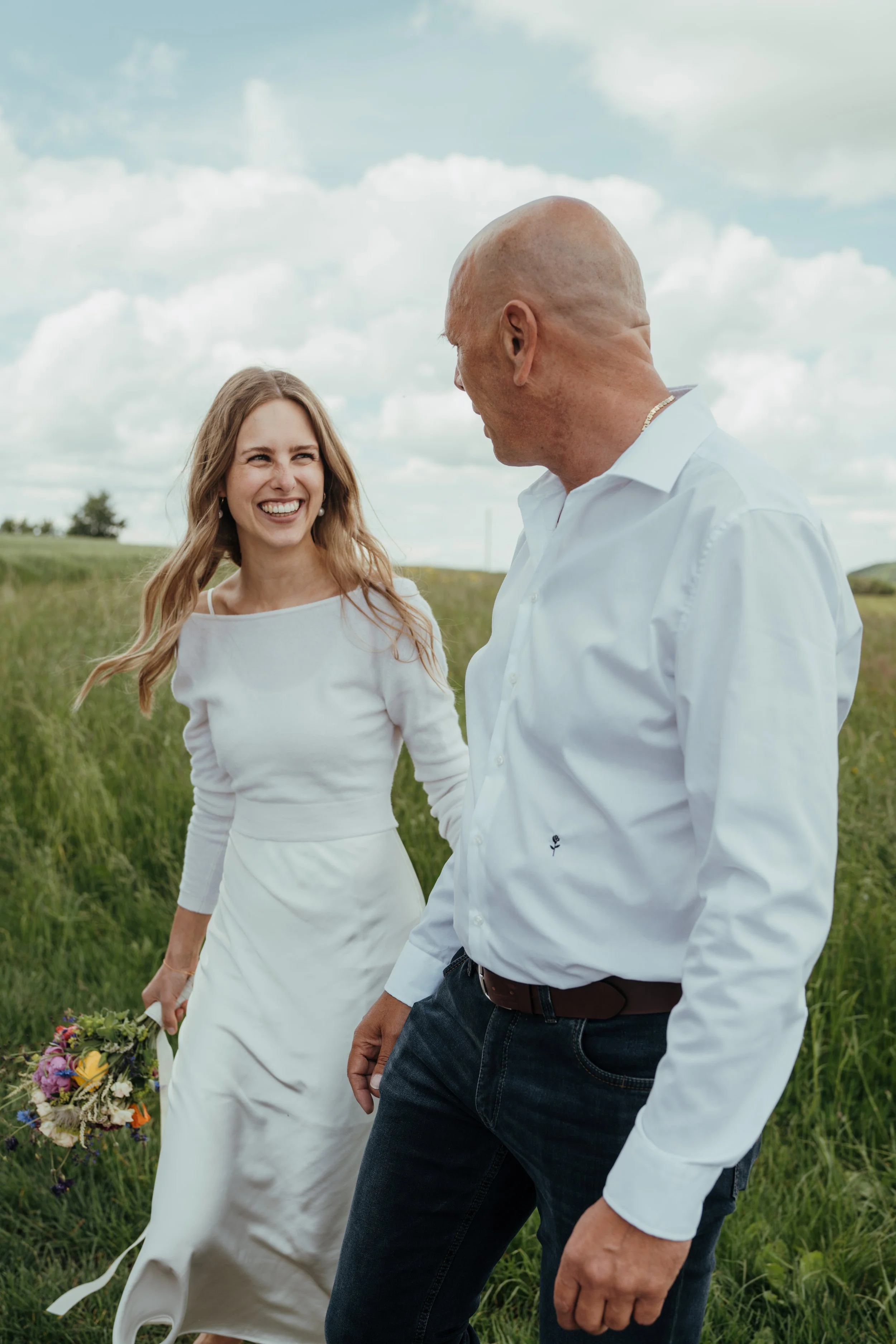 Ein Mädchen in einem weißen Kleid hält einen Blumenstrauß, während ein Mann in einem weißen Hemd und blauer Jeans mit einem anderen Mann spricht, in einem grünen Feld bei bewölktem Himmel.