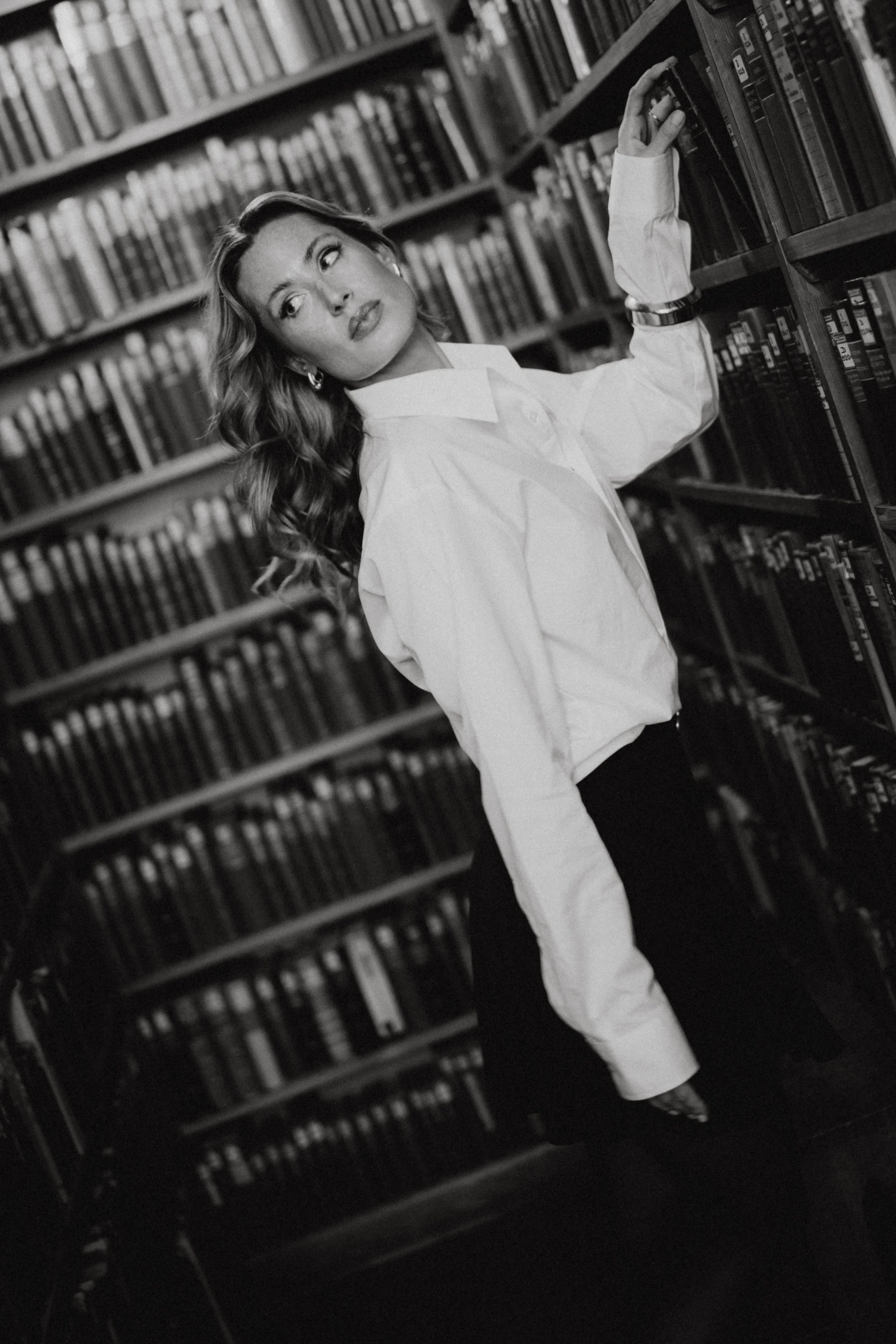 Schwarz-weiß-Foto einer jungen Frau in einer Bibliothek, die Bücherregale durchstöbert und an einem Regal zieht.