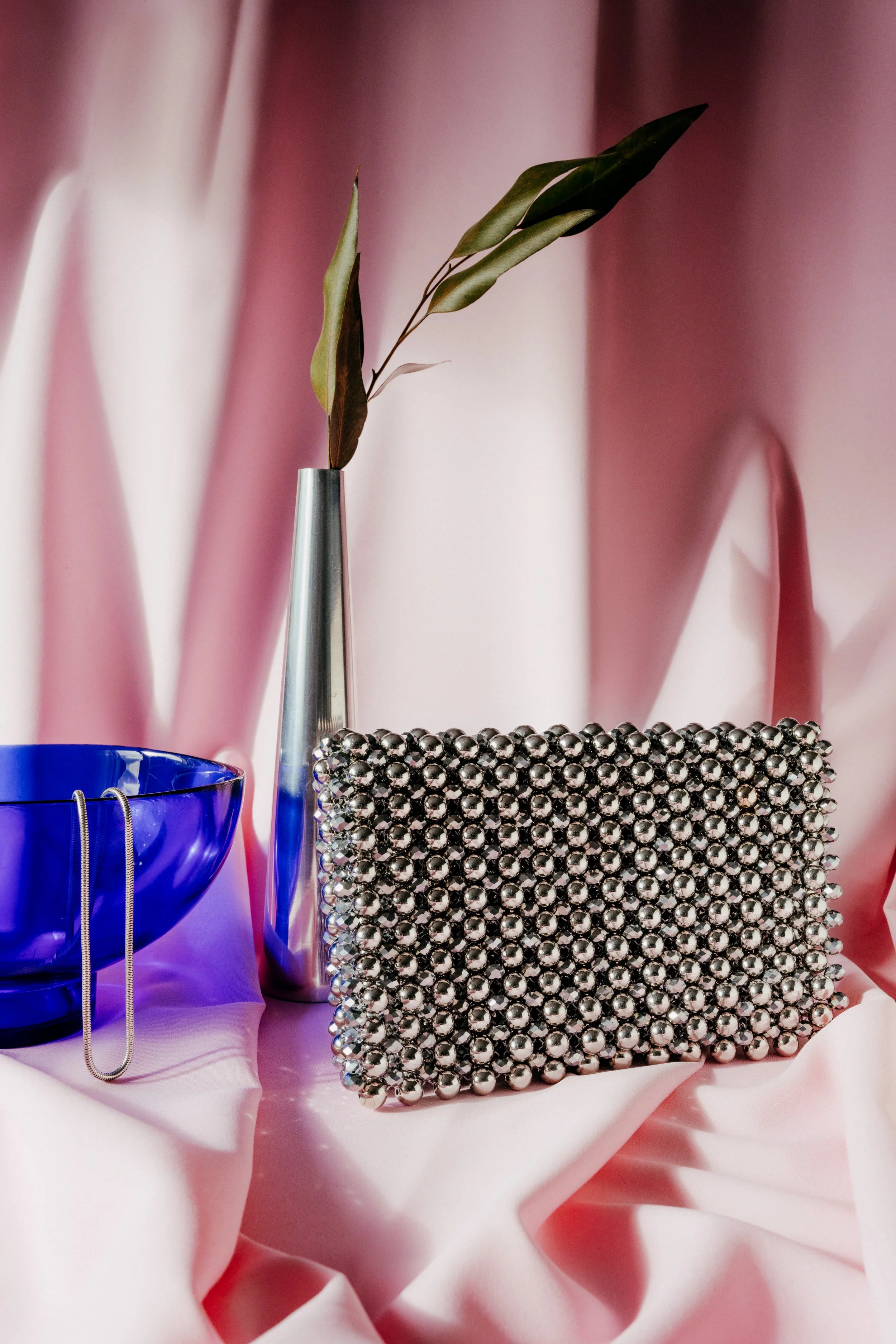 Ein silberner Würfel aus Metallkugeln, eine blaue Glasschale mit silberner Kette, ein Blumentopf mit Pflanze auf rosa Satin-Hintergrund.