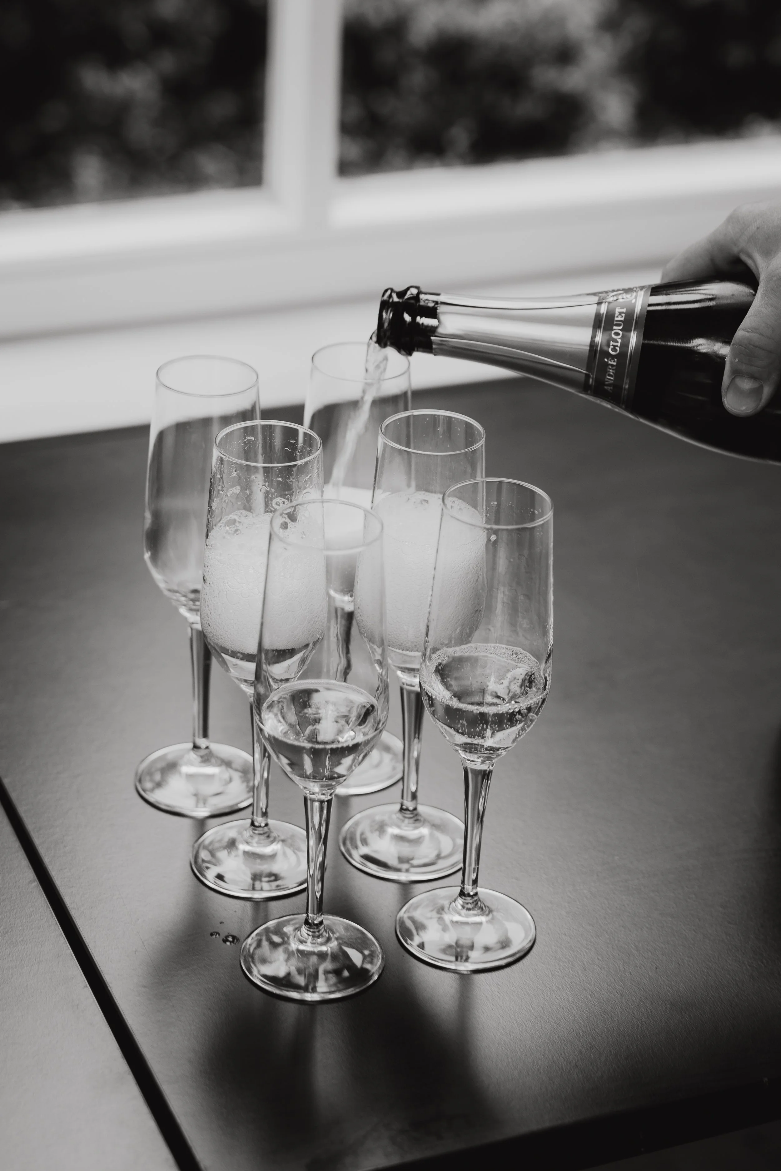 Ein Hand, die eine Flasche Champagner in ein Glas einschenkt, mehrere leere Champagnergläser stehen auf einem Tisch.
