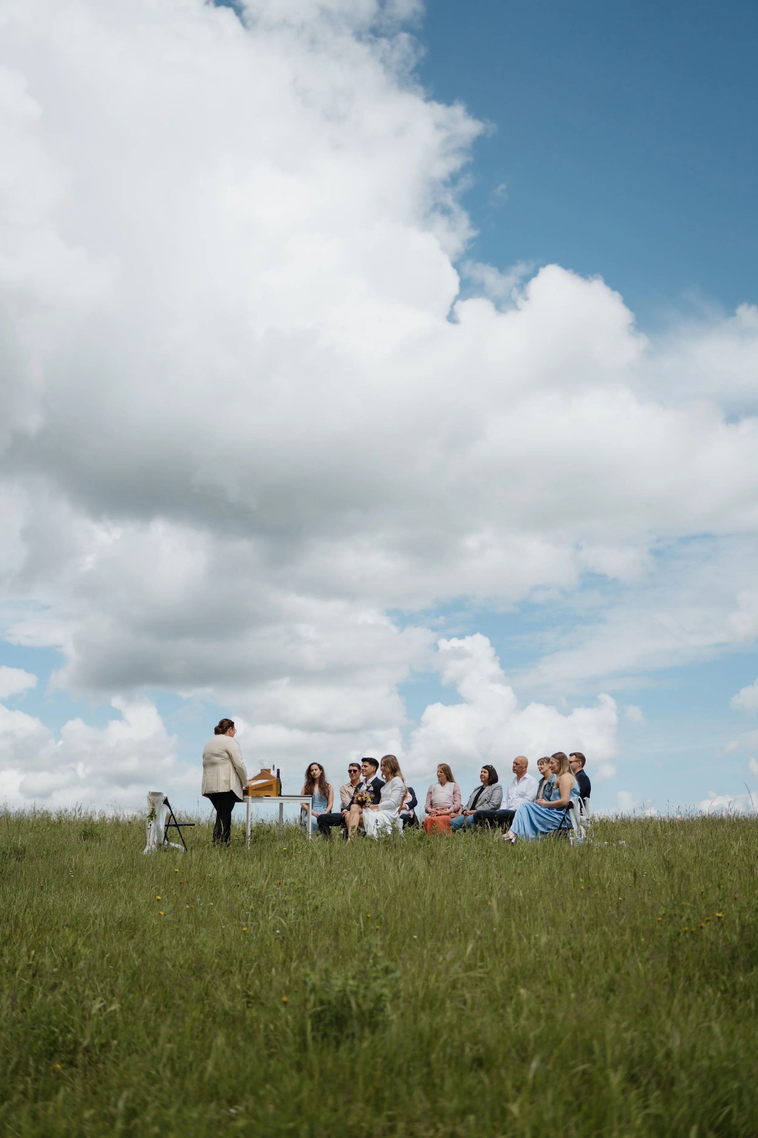 Eine Gruppe von Menschen sitzt im Freien auf Stühlen in einer großen Wiese bei einer Zeremonie, während eine Person an einem Klavier spielt. Der Himmel ist überwiegend bewölkt mit einigen blauen Bereichen.