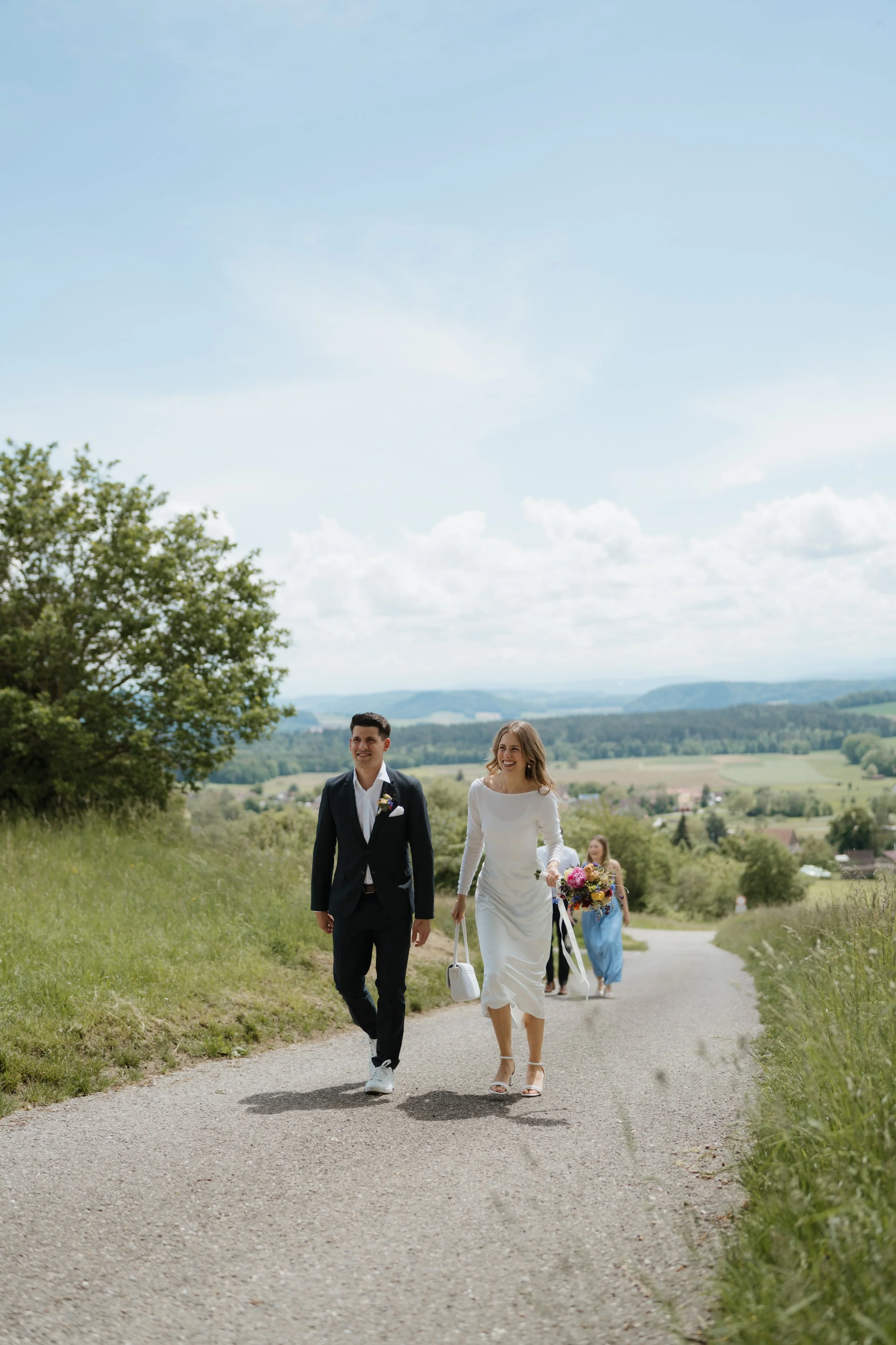 Ein frisch verheiratetes Paar läuft auf einer Landstraße in der Natur. Der Mann trägt einen schwarzen Anzug und die Frau ein weißes Hochzeitskleid und trägt einen Blumenstrauß.