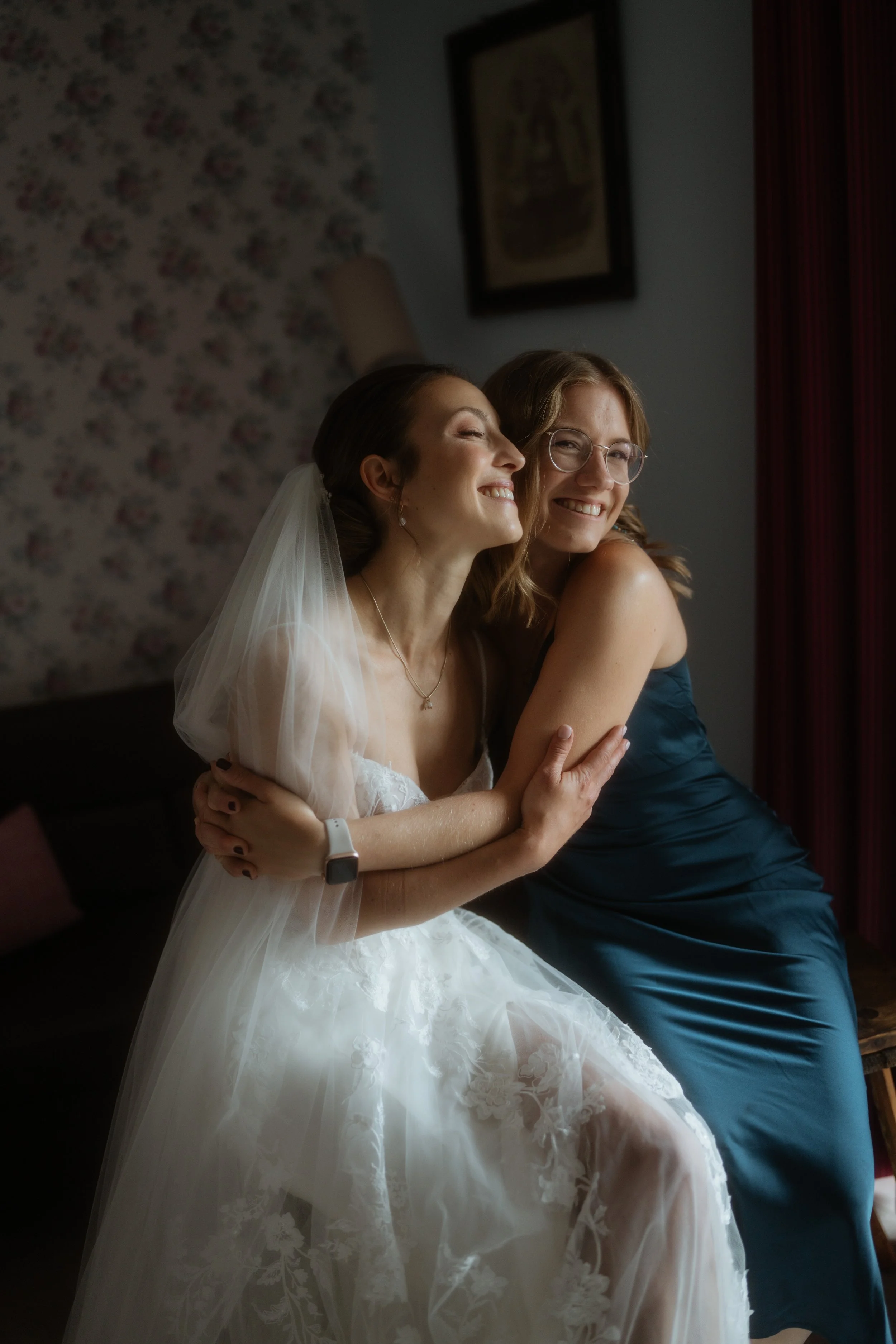 Zwei Frauen, eine im Brautkleid und die andere in einem blauen Kleid, umarmend und lachend in einem Zimmer mit dunklem Hintergrund und Blumengestaltung.