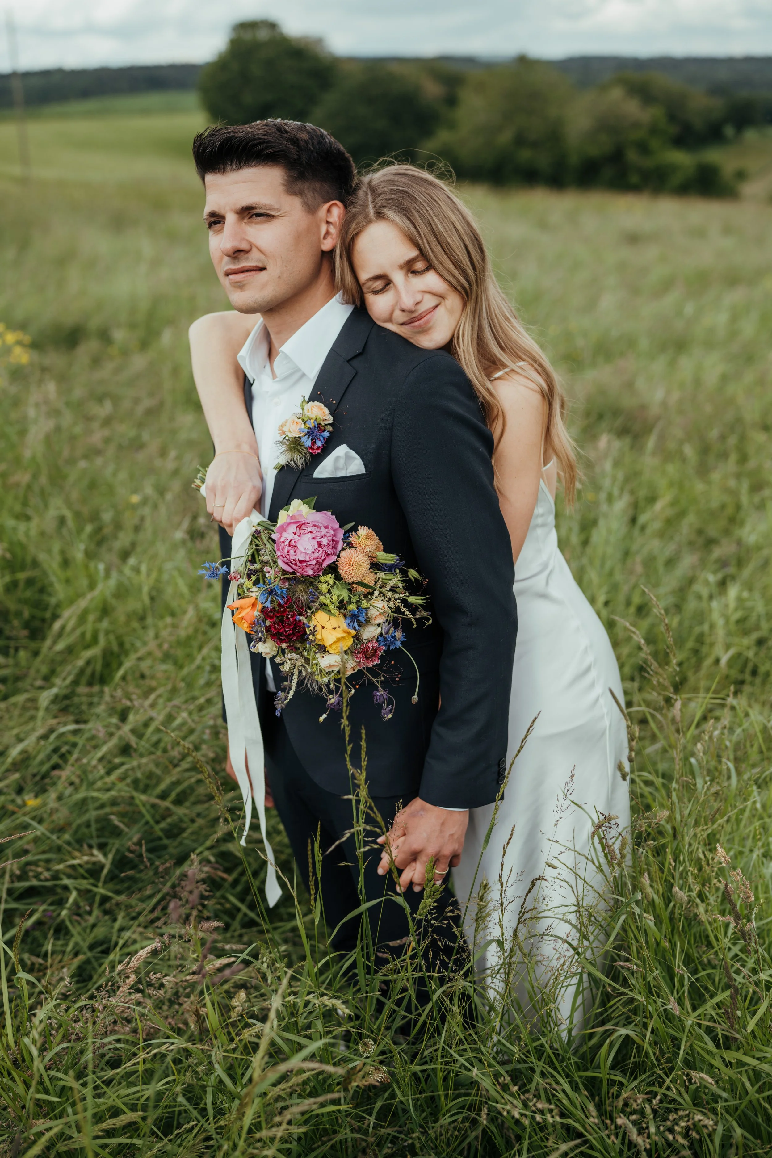 Ein glückliches Paar, ein Mann in Anzug mit Blumen an der Jacke, eine Frau in einem weißen Kleid, umarmt sich in einer grünen Wiese mit Bäumen im Hintergrund.