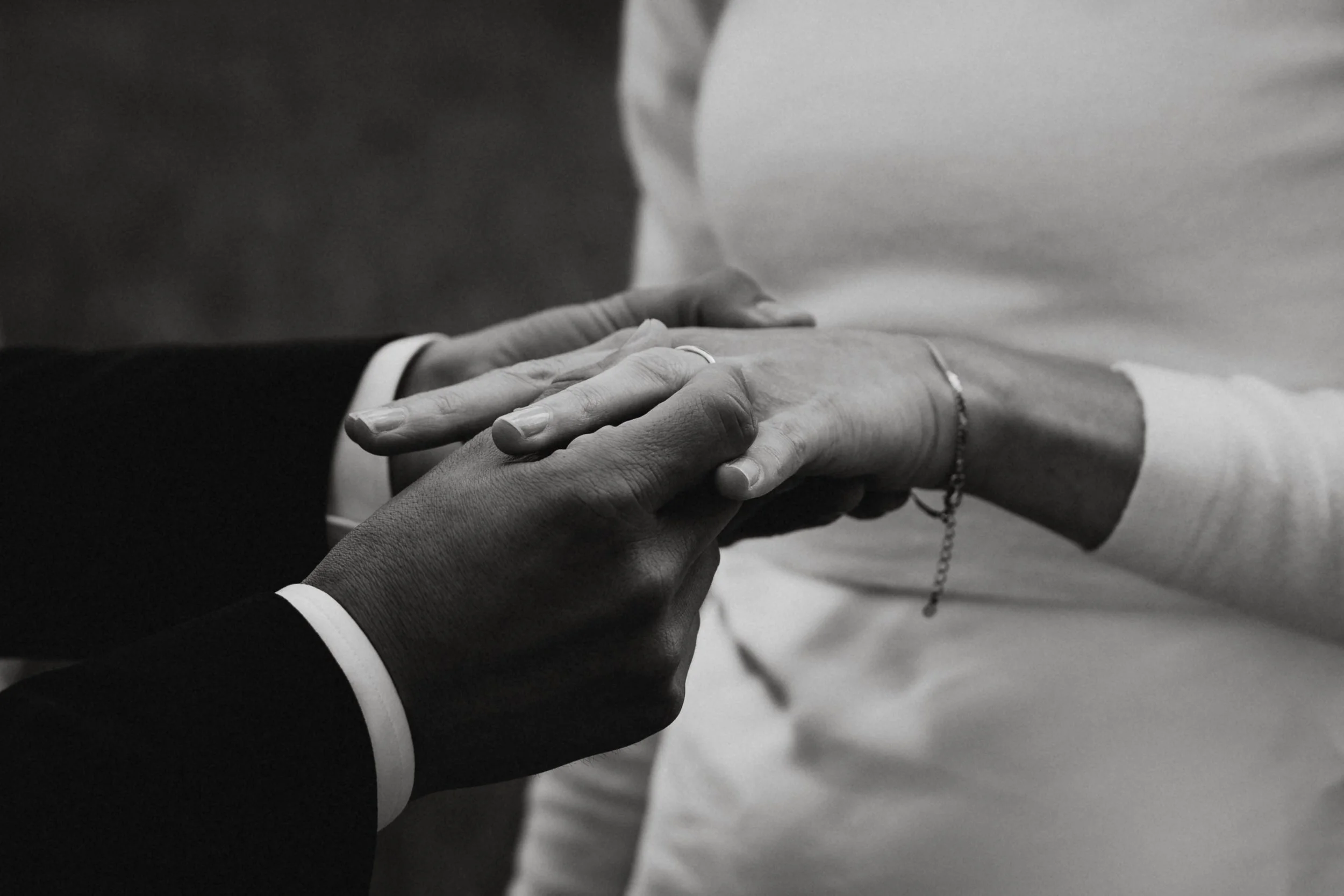 Zwei Menschen halten sich an den Händen, eine Person trägt einen Ring, die andere Person trägt eine Armbanduhr und eine Ketten. Die Szene ist schwarz-weiß.