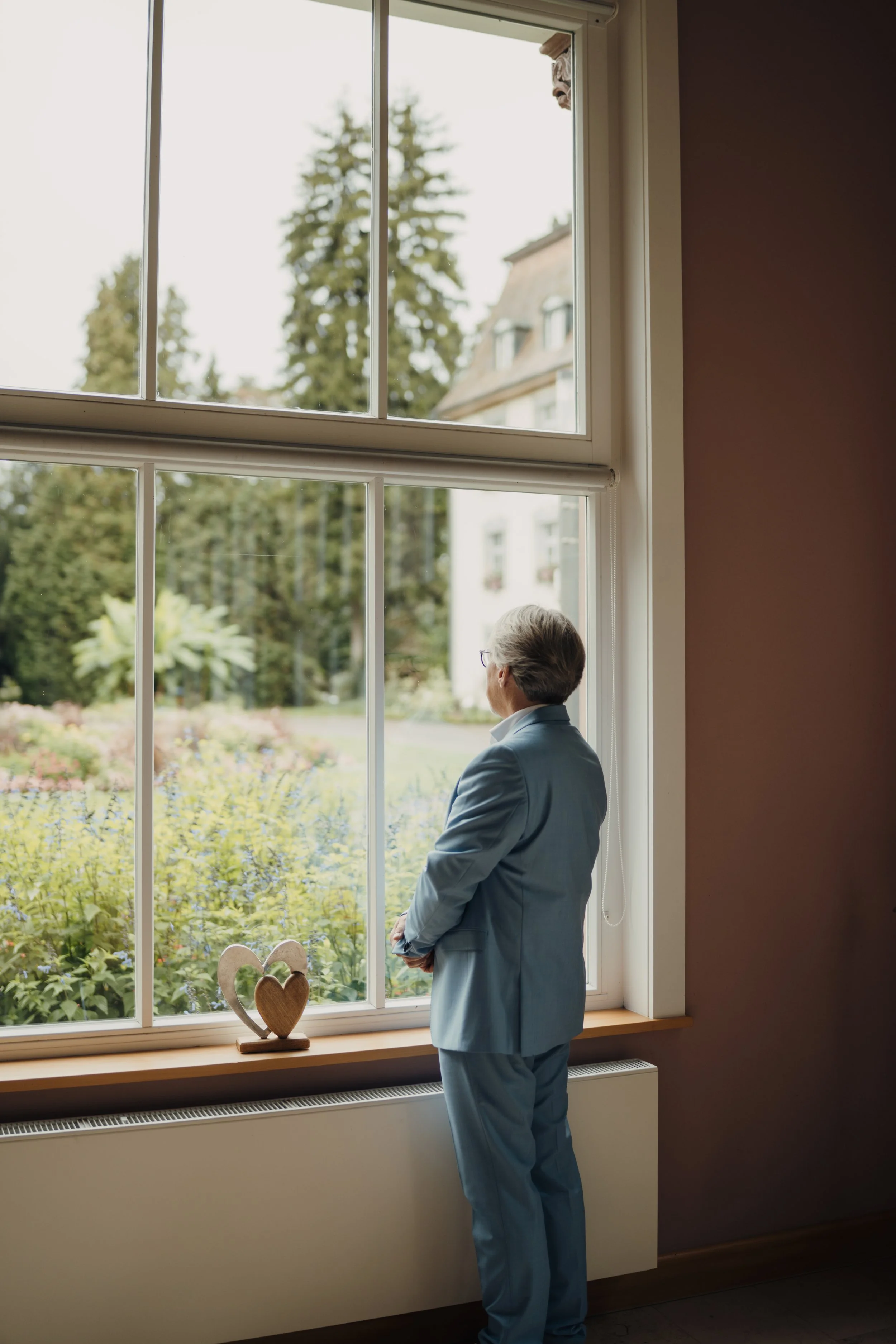 Ein älterer Mann in einem hellblauen Anzug steht vor einem großen Fenster und schaut nach draußen auf einen Garten mit Büschen und Bäumen. Auf der Fensterbank steht eine Holz-Deko in Herzform.