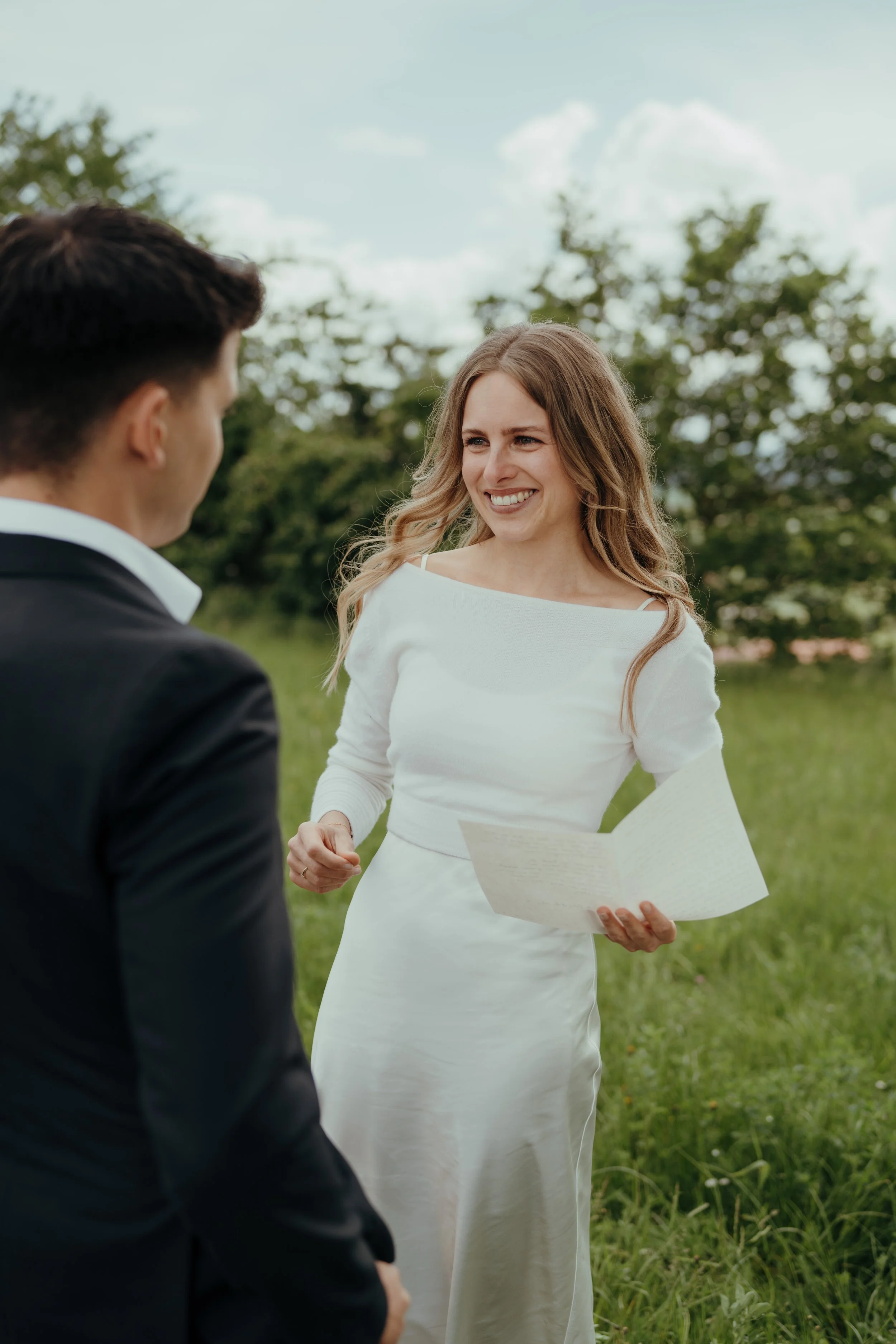 Eine Frau in einem weißen Kleid hält ein Dokument und lächelt einen Mann in einem schwarzen Anzug an, während sie im Freien auf einer Wiese stehen, umgeben von Bäumen und einem Himmel mit Wolken.