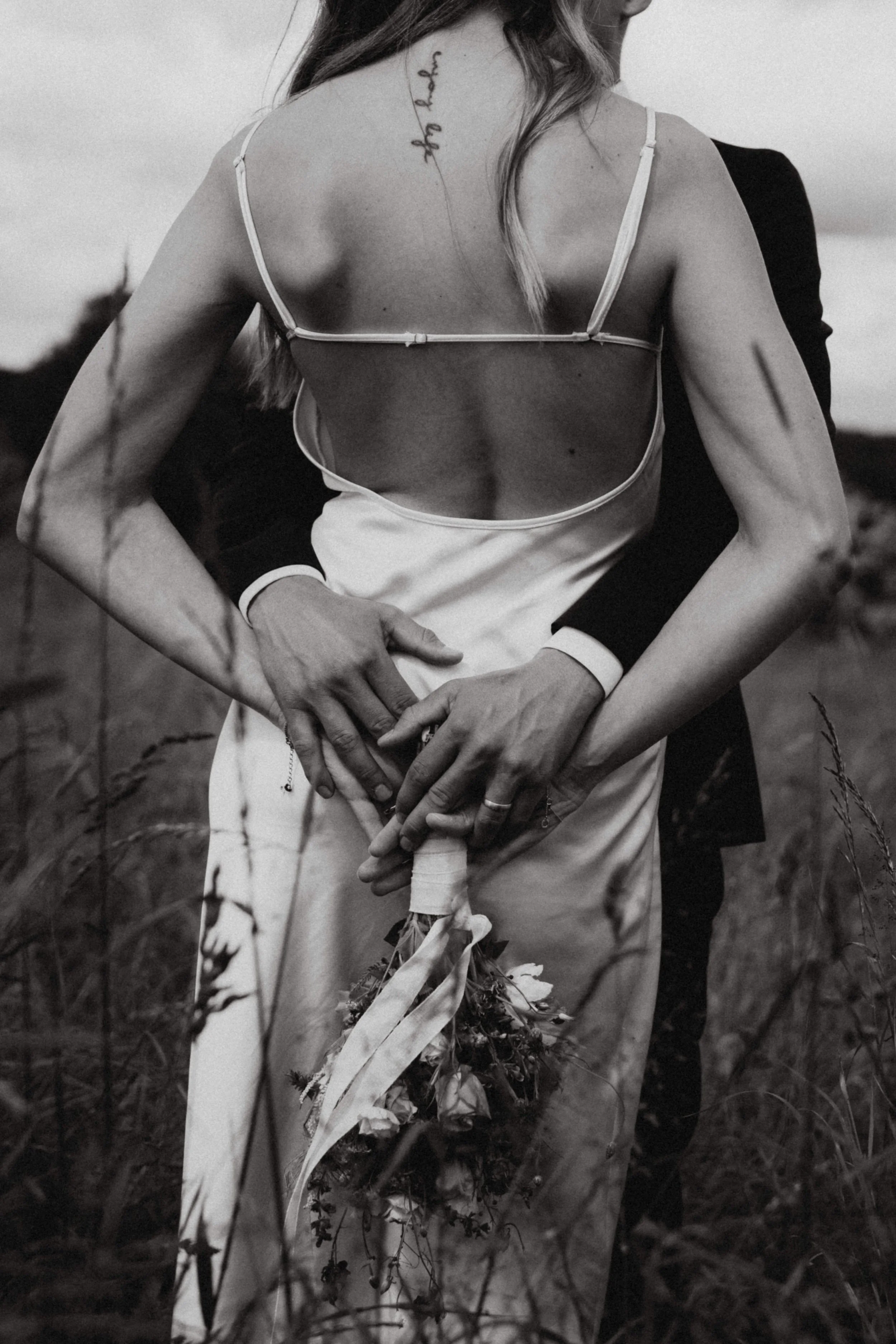 Schwarz-weiß-Foto einer Frau, die in einem Feld steht, trägt ein ärmelloses Kleid mit Trägern und hält Blumen in den Händen. Rückenansicht, Tattoo auf der Schulter, lange Haare.