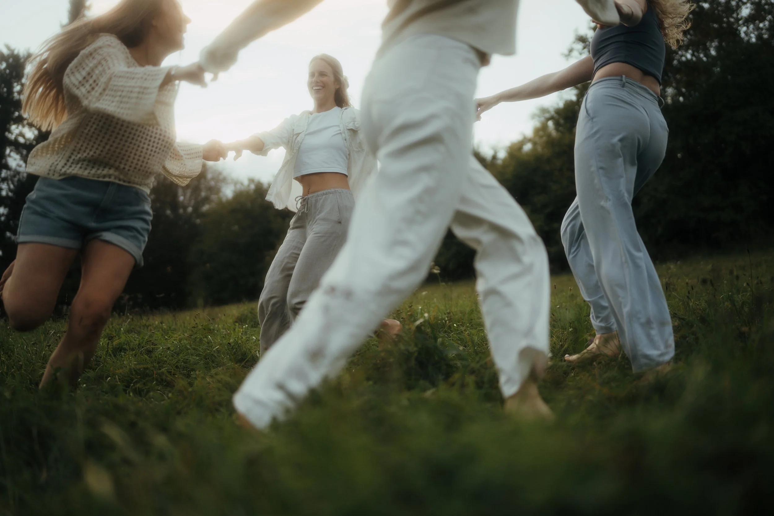 Sechs junge Menschen drehen sich im Kreis, halten sich an den Händen und lachen, draußen in einem grünen Gebiet mit Bäumen im Hintergrund, bei Sonnenuntergang oder -aufgang.