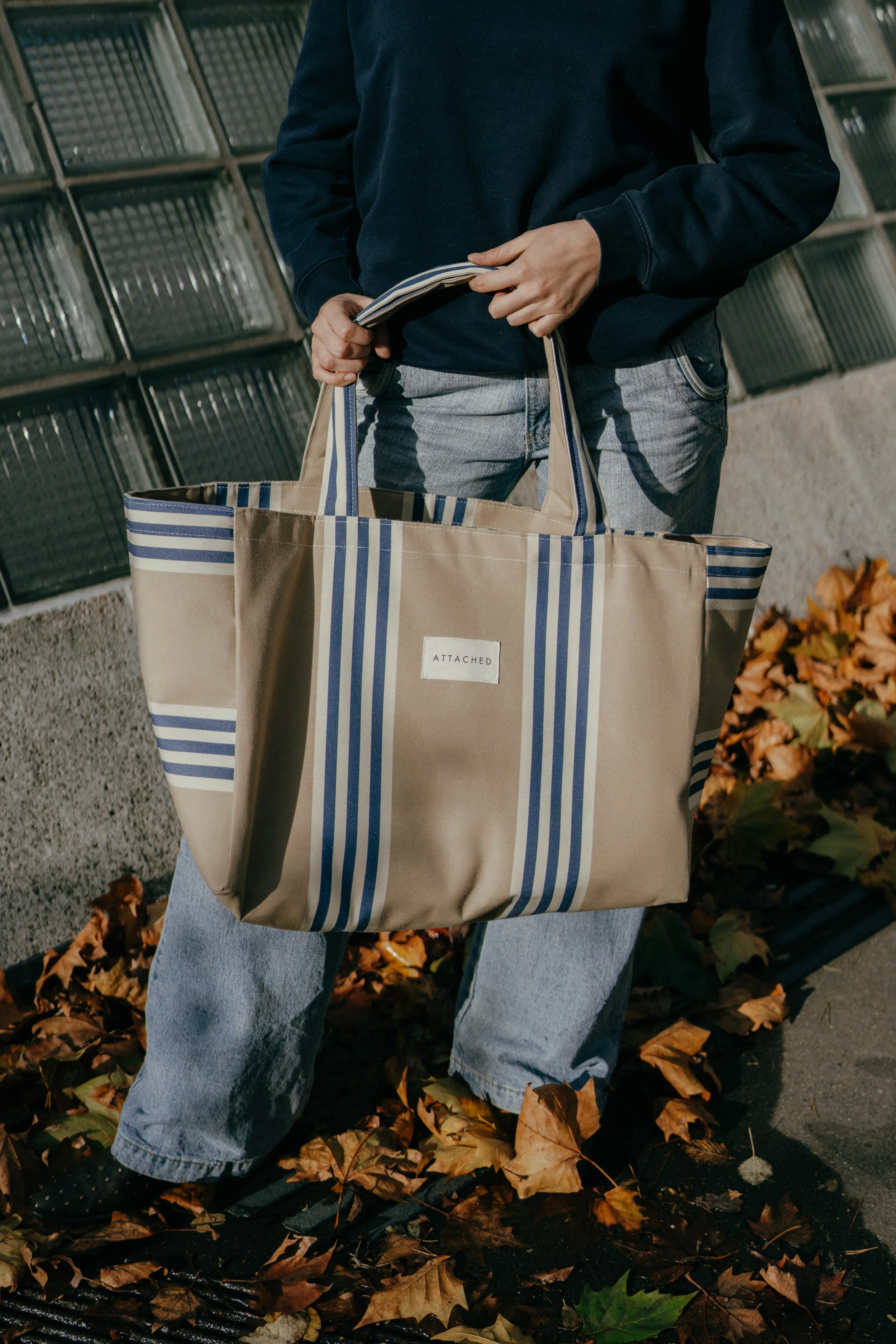 Person mit blauen Jeans, schwarzem Pullover und Tragetasche aus Stoff mit Streifenmuster, umgeben von Herbstblättern.