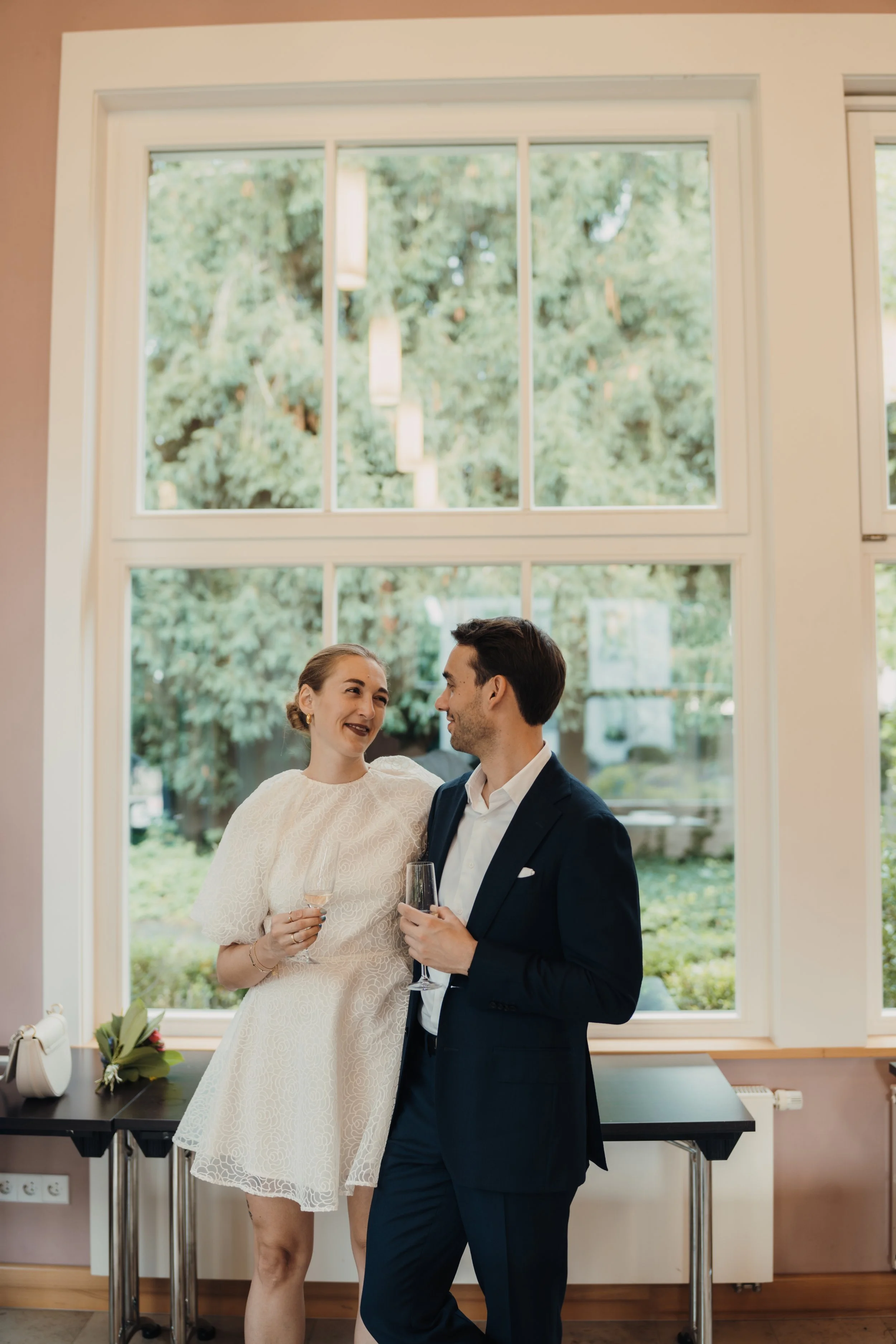 Ein Mann und eine Frau bei einer Feier, die in einem Raum mit großen Fenstern und Blick ins Grüne stehen. Die Frau trägt ein weißes Kleid und hält ein Glas Sekt, der Mann trägt einen dunklen Anzug.