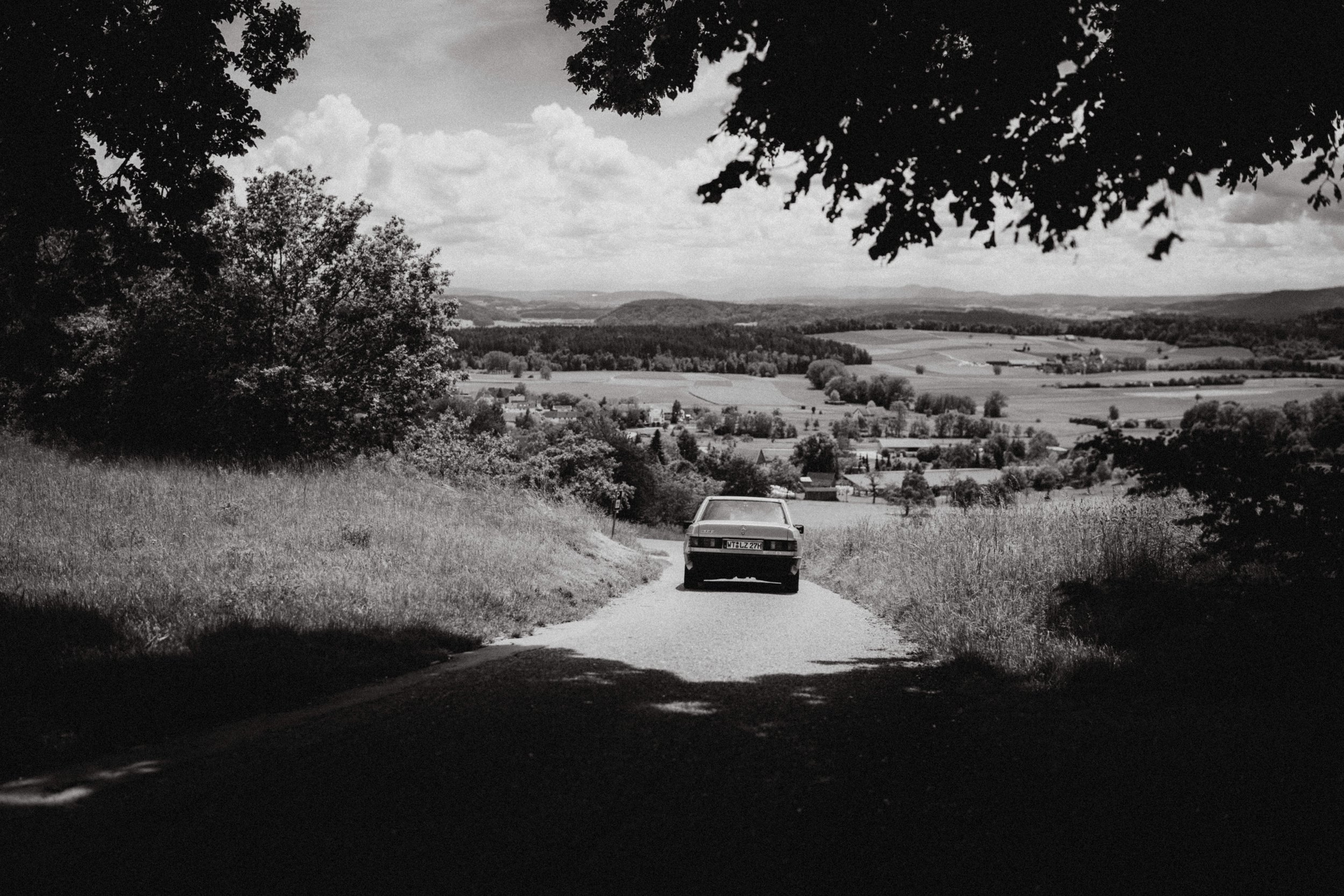 Schwarzes und weißes Foto einer Landstraße mit einem alten Auto, umgeben von Bäumen und weiter in die Landschaft mit Feldern und Hügeln im Hintergrund.