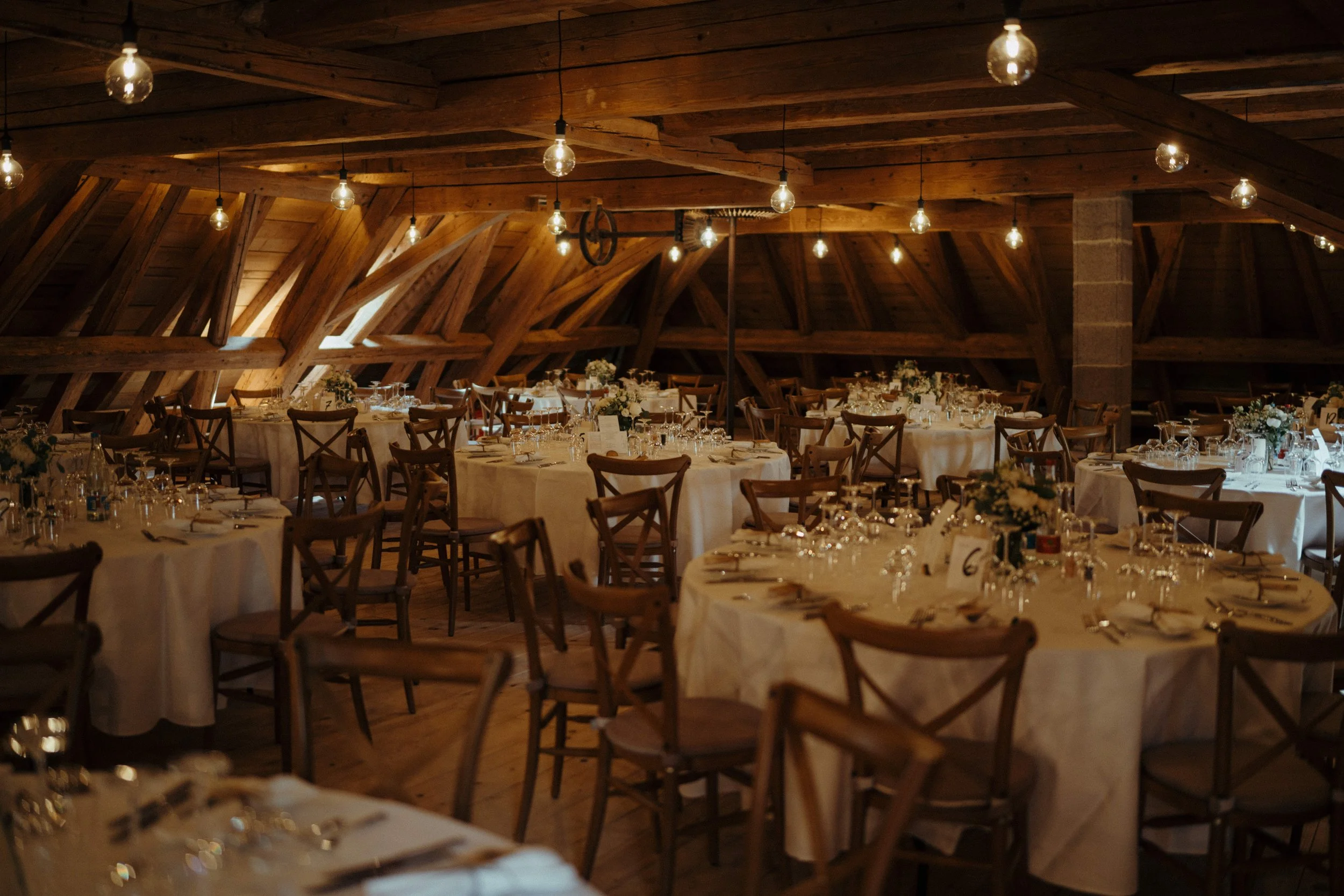 Ein festlich gedeckter Veranstaltungsraum mit runden Tischen, weißen Tischdecken, Kerzen, Blumenarrangements und Wein- oder Sektgläsern, dekoriert für eine Feier in einem rustikalen Raum mit Holzdecke und sichtbaren Holzbalken.
