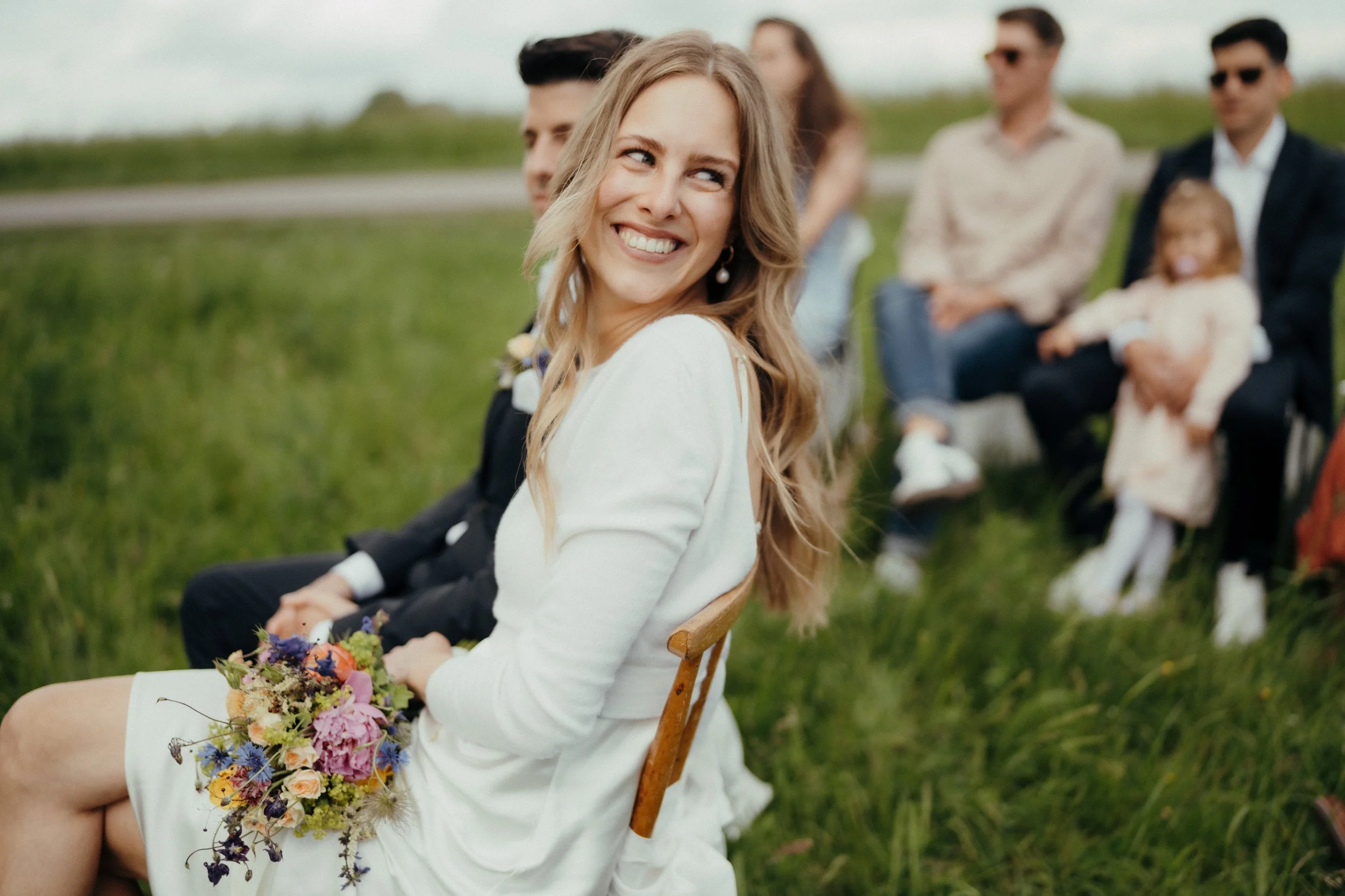 Junge Frau sitzt im weißen Kleid mit Blumenstrauß auf dem Schoß, lächelnd, im Freien, umgeben von anderen Menschen, die im Hintergrund sitzen, bei Sonnenschein.