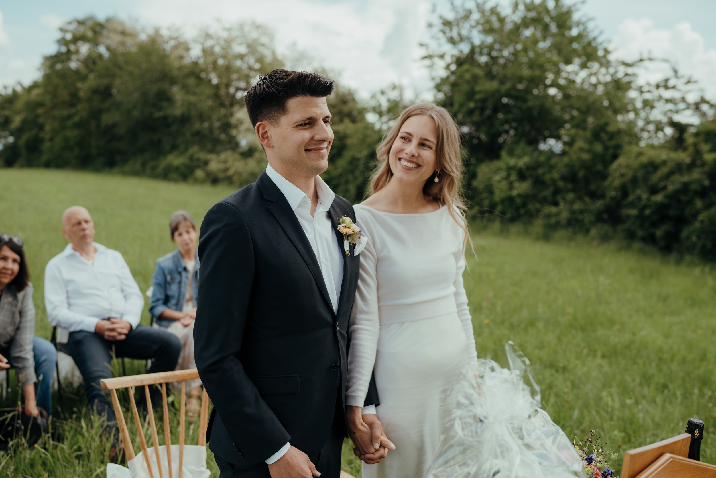 Ein Brautpaar bei einer Hochzeit im Freien, Hand in Hand, umgeben von Freunden und Familie, bei schönem Wetter mit Wolken am Himmel.
