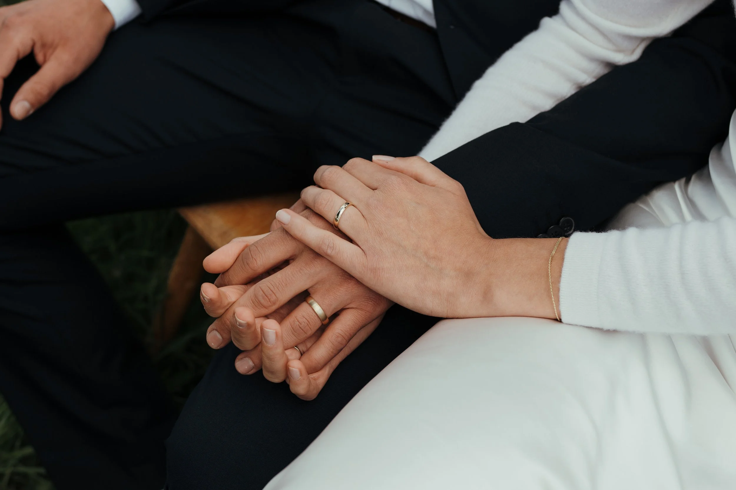 Zwei Menschen halten sich an den Händen, die eine trägt einen Ehering, die andere eine schmale goldene Kette. Die Personen tragen formelle Kleidung, eine in schwarz und die andere in weiß.