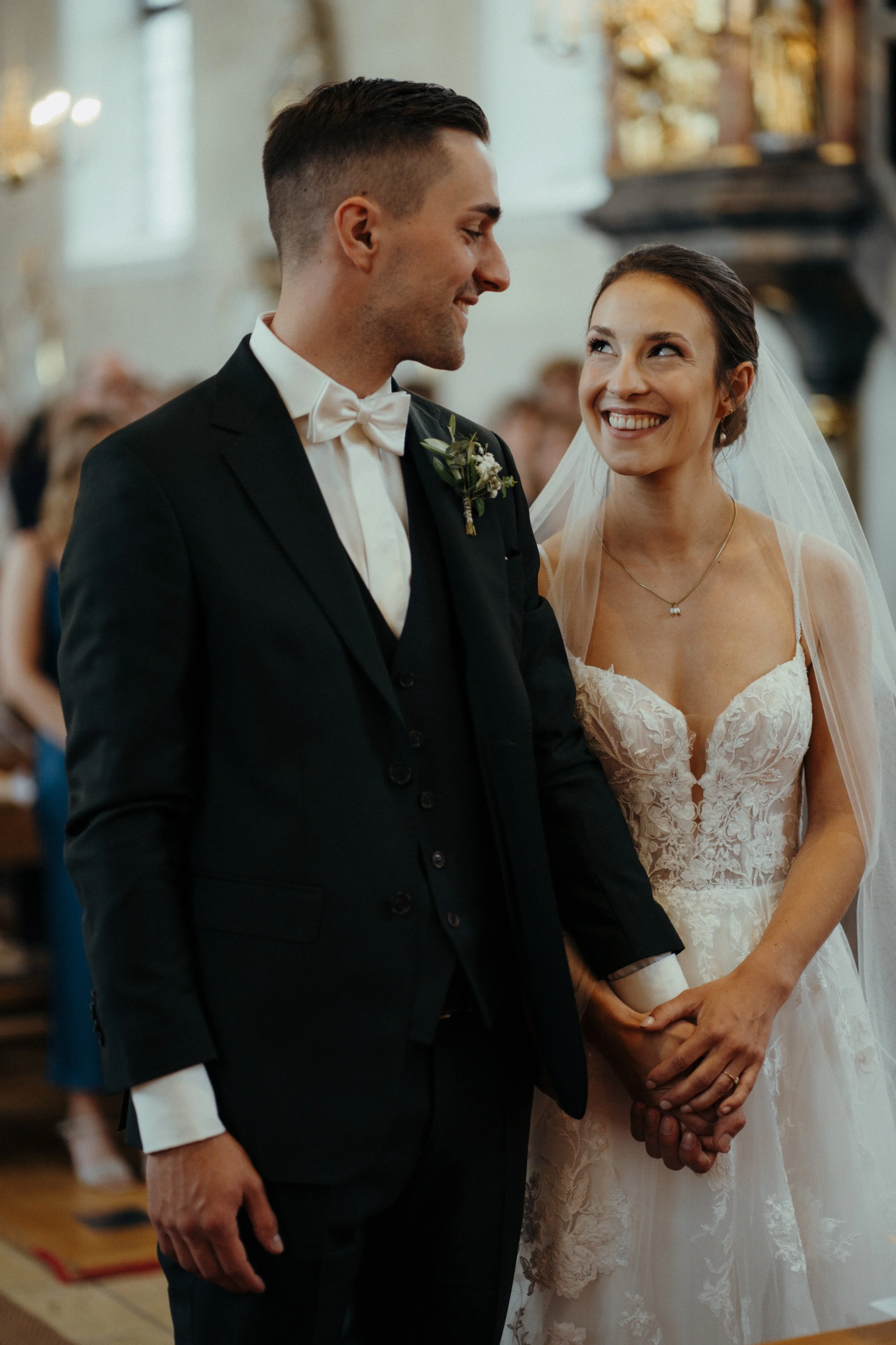 Ein Pärchen bei ihrer Hochzeit in einer Kirche, das Händchen hält und sich liebevoll ansieht.