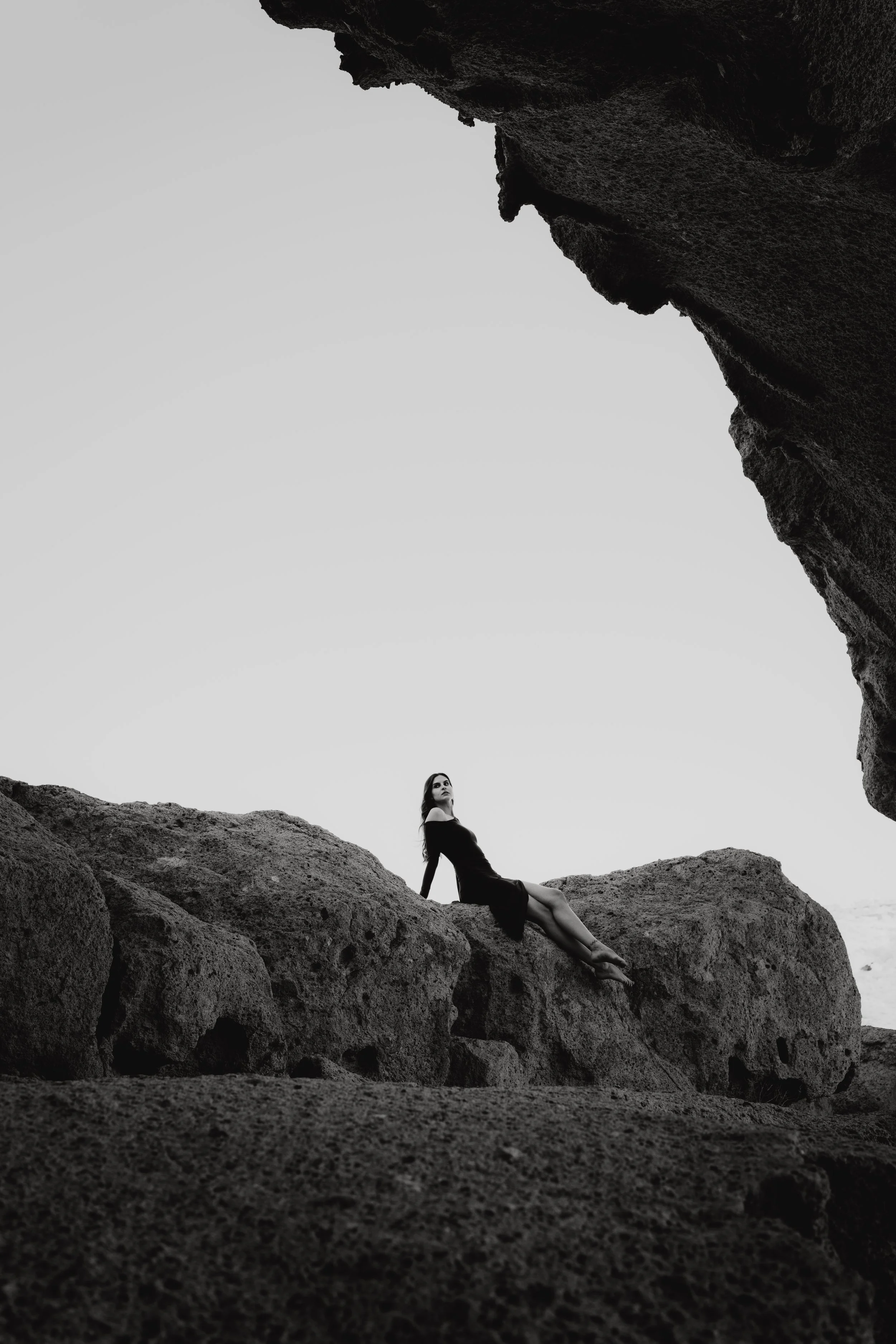 Eine Frau in einem schwarzen Kleid sitzt auf einem großen Felsen an einer Küste, umgeben von weiteren Felsen, bei Sonnenuntergang oder Sonnenaufgang, im schwarz-weiß Stil