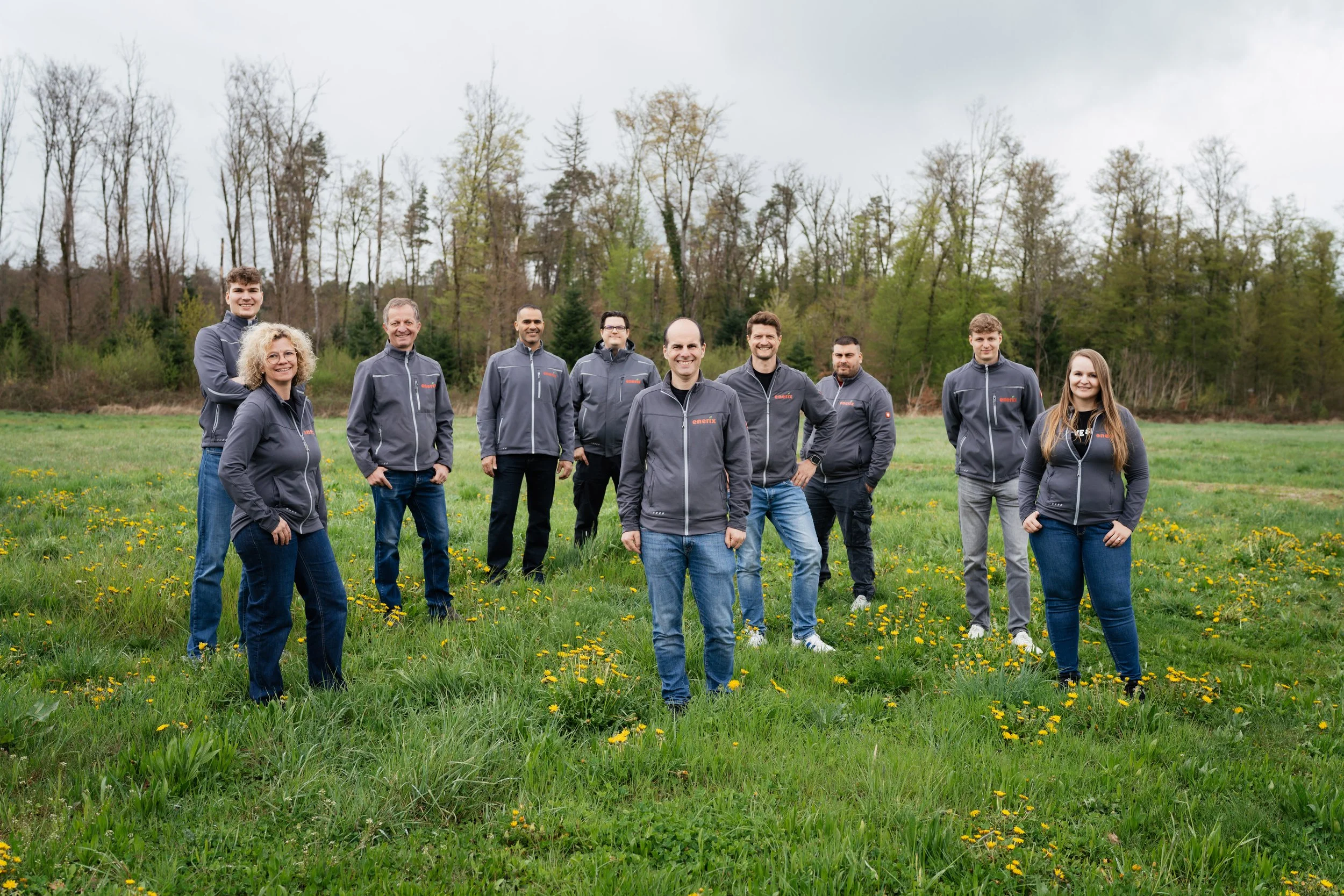 Eine Gruppe von acht Personen steht lachend auf einer grünen Wiese mit kleinen gelben Blumen, im Hintergrund Bäume und ein bewölkter Himmel.