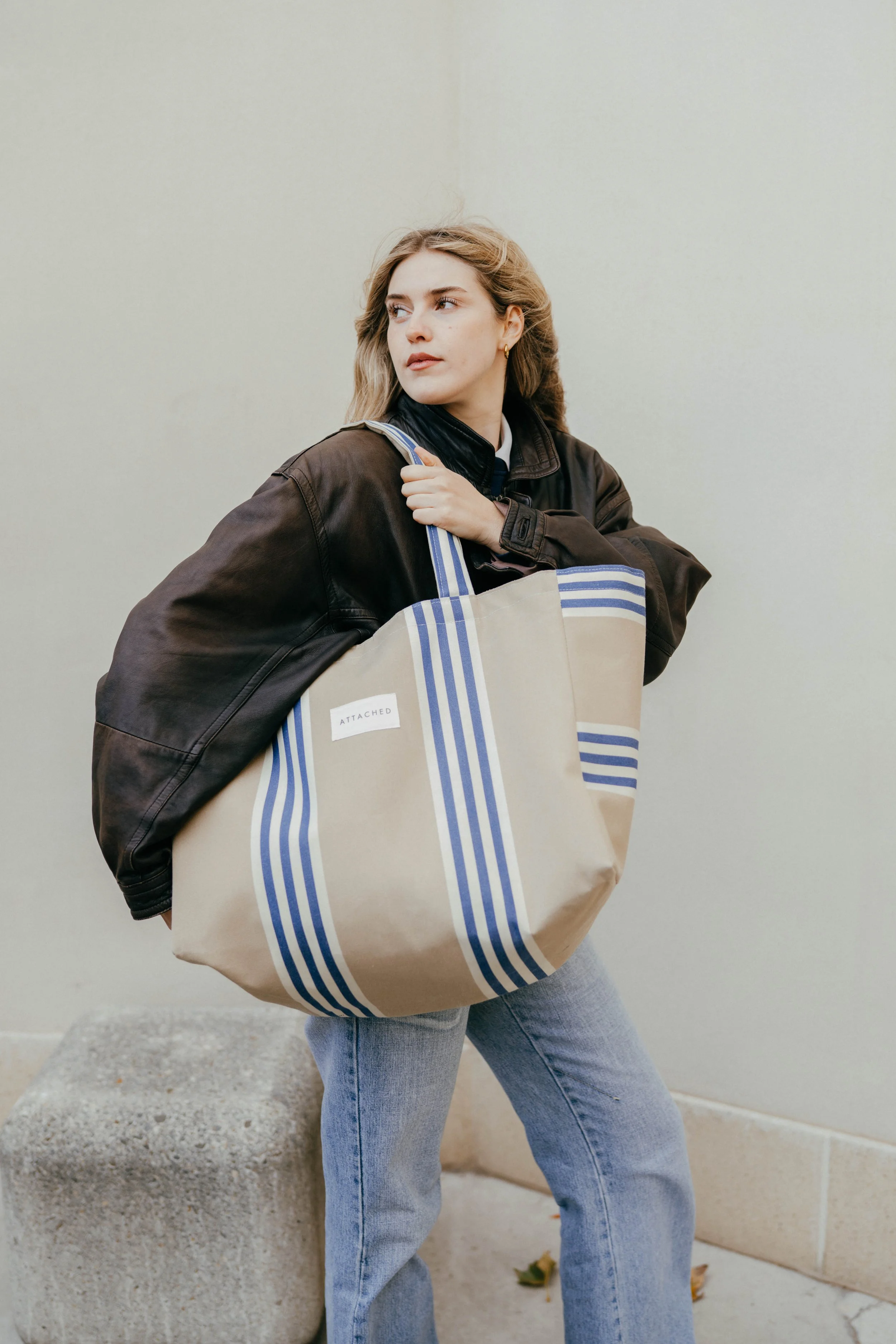 Junge Frau mit Lederjacke und Jeans, die eine große beige Einkaufstasche mit blauen Streifen trägt, steht neben einem Betonblock vor einer hellen Wand.