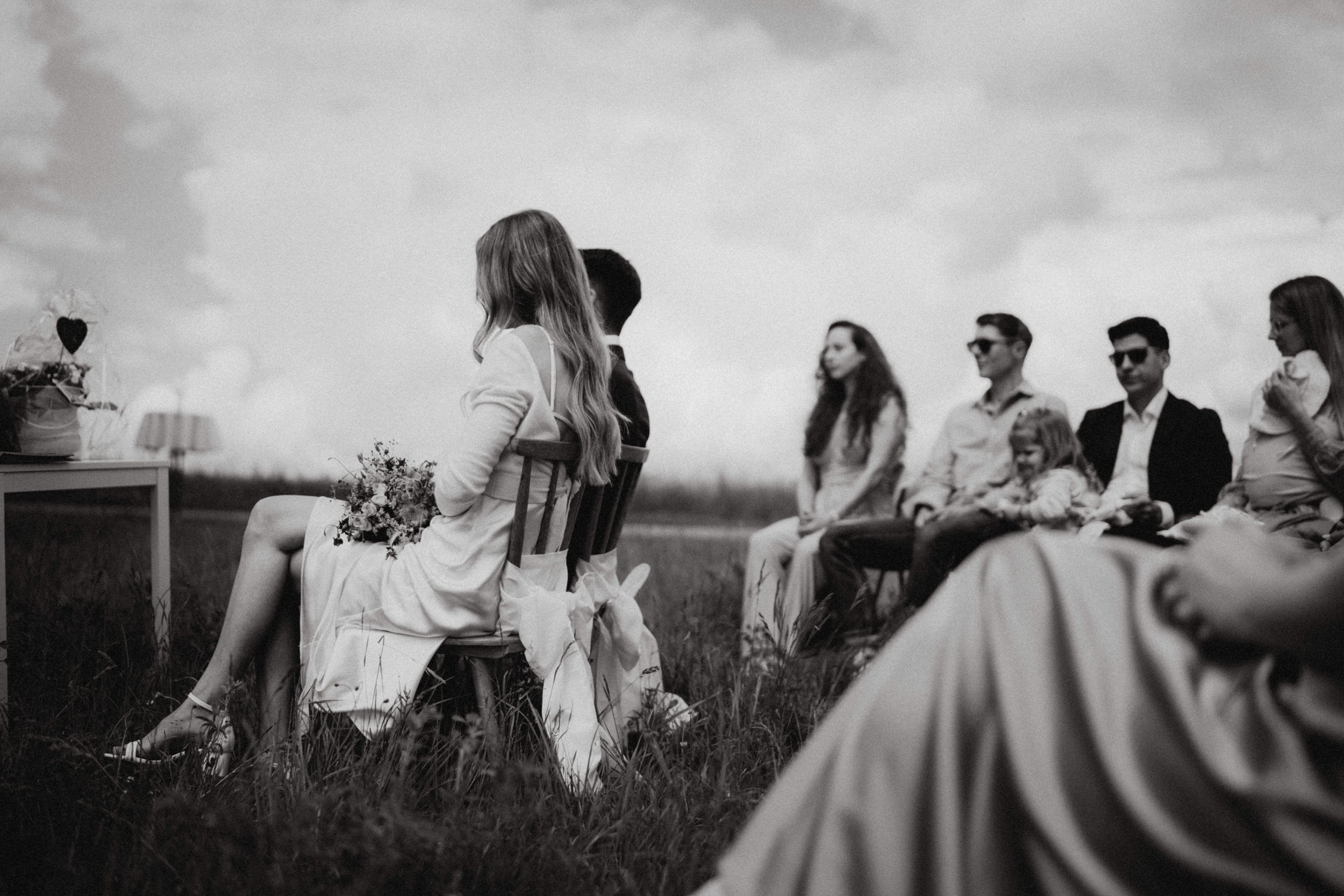 Schwarz-Weiß-Foto einer Gruppe von Menschen, die im Freien sitzen, vermutlich bei einer Hochzeit oder Feier, mit Fokus auf eine sitzende Frau mit langen, gelockten Haaren, die einen Blumenstrauß hält.