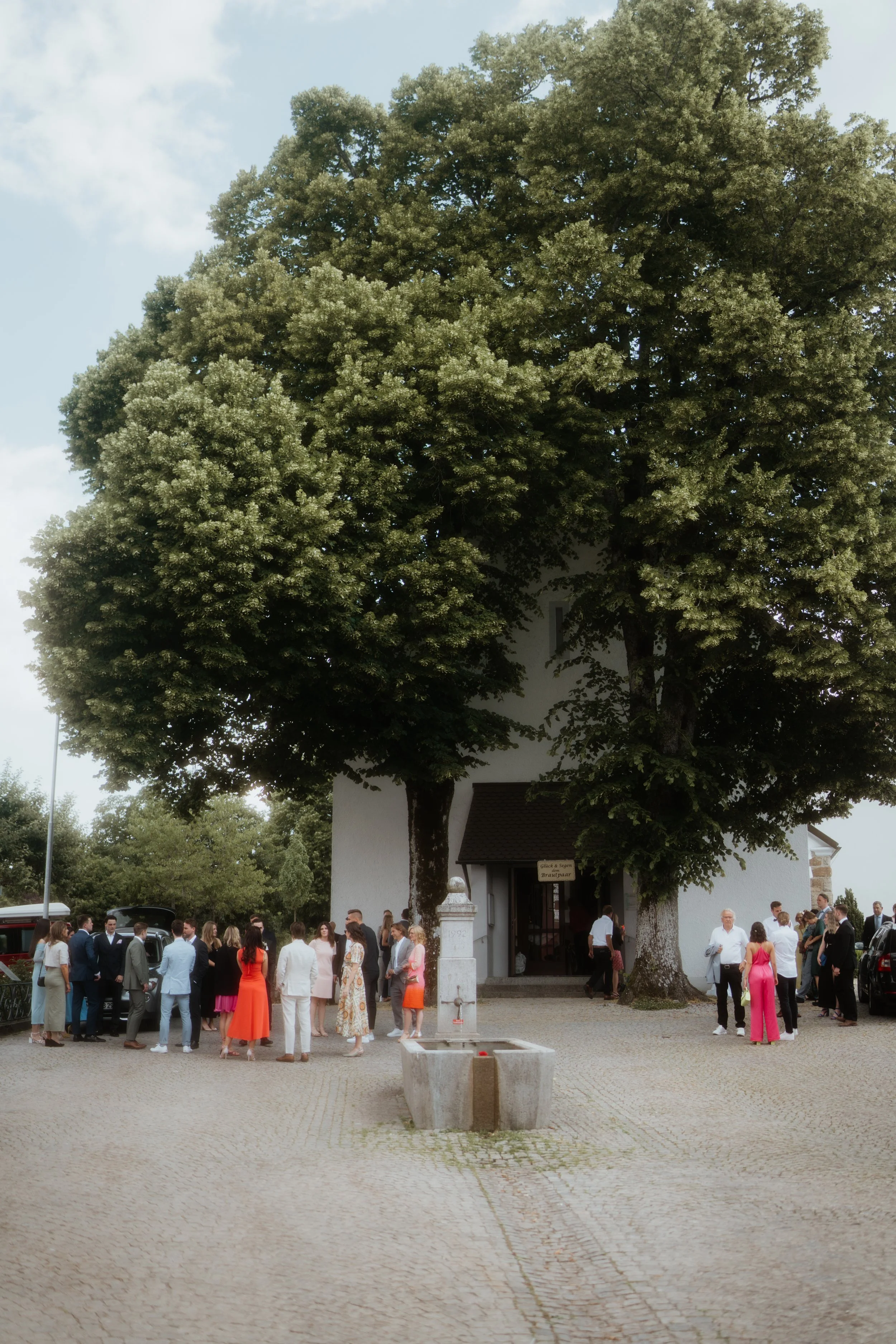 Menschen in festlicher Kleidung versammeln sich vor einem weißen Gebäude mit großen Bäumen im Hintergrund bei einer Feier oder Hochzeit auf dem Platz vor dem Gebäude.