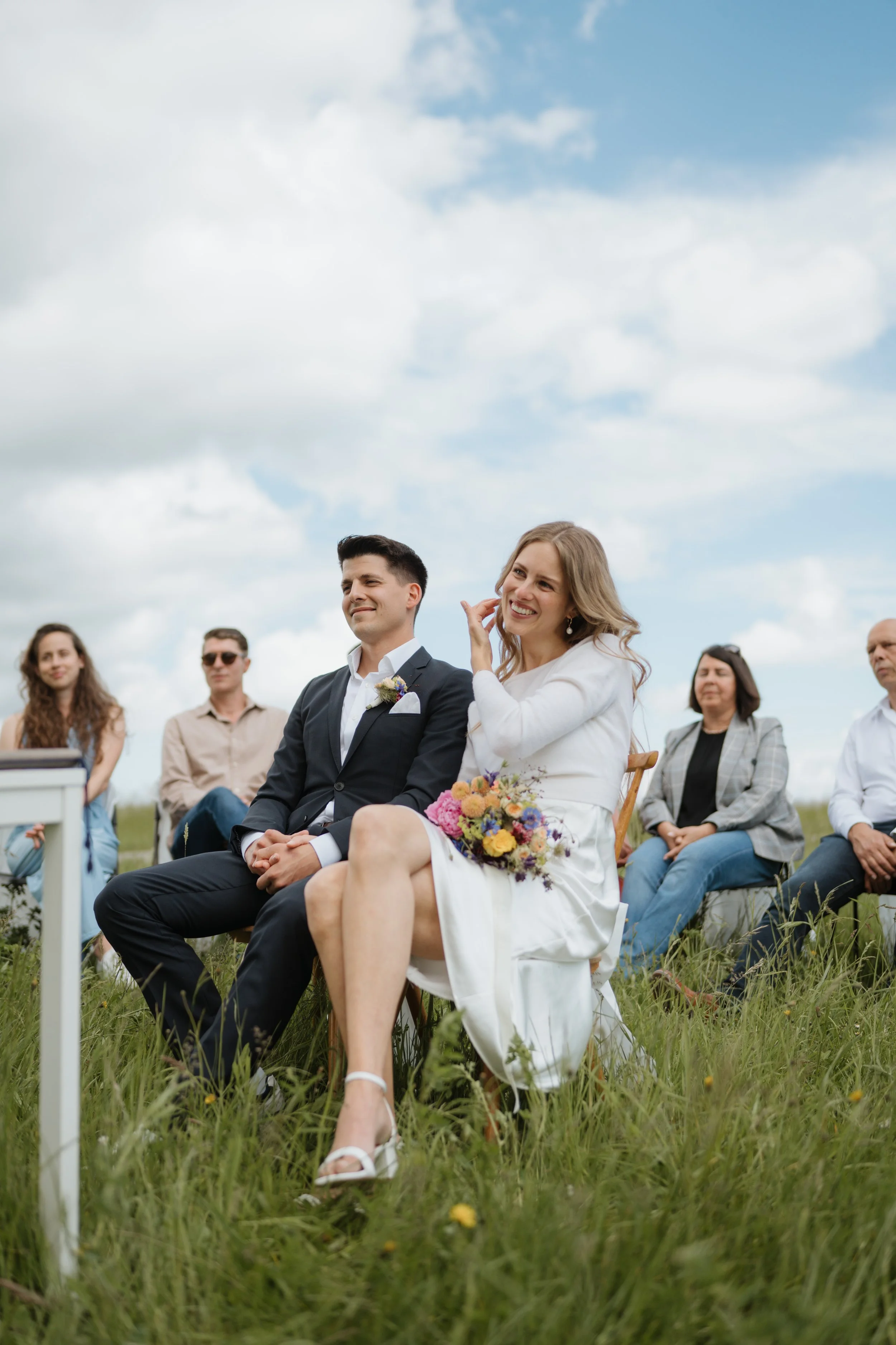 Ein Brautpaar bei einer Freilufthochzeit, sitzend auf Stühlen im Gras, umgeben von Freunden und Familie, im Himmel blauer Himmel mit Wolken.