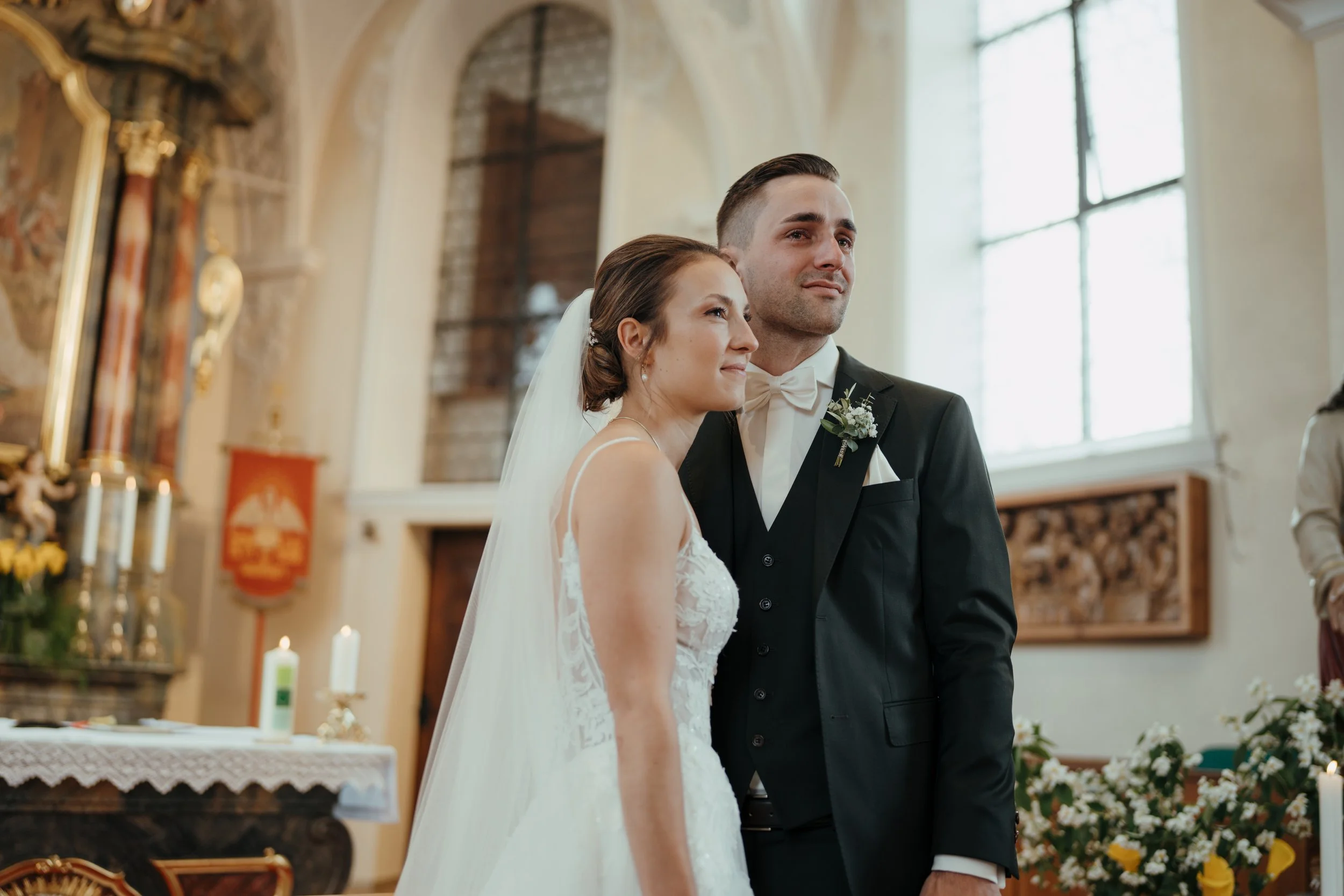 Ein Brautpaar bei einer Trauung in einer Kirche, die Braut trägt ein weißes Kleid und der Bräutigam einen schwarzen Anzug mit Fliege. Sie stehen nebeneinander vor einem Altar.