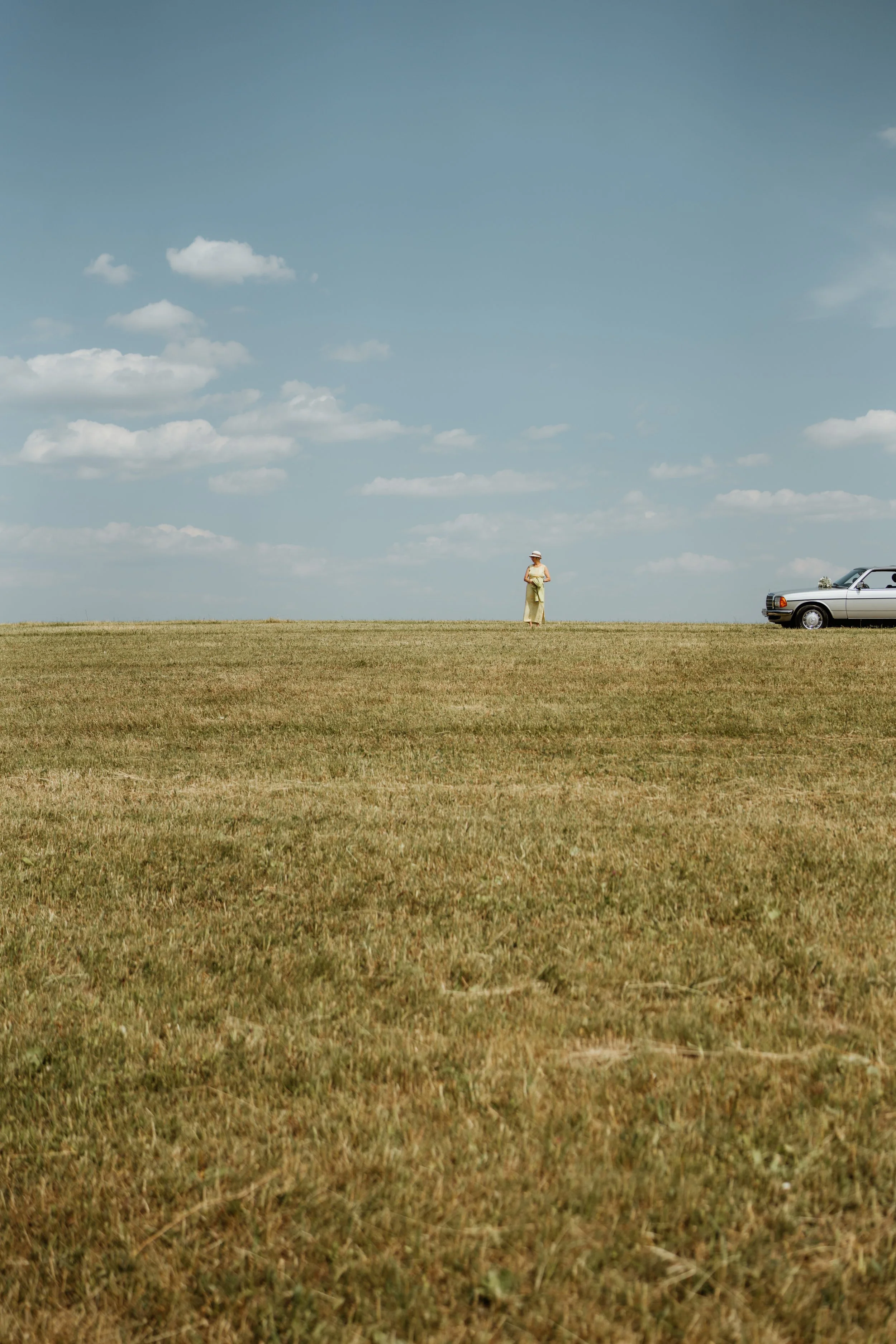 Eine Frau steht in einem offenen, grasbewachsenen Feld unter einem klaren Himmel mit einigen Wolken. Auf der rechten Seite ist ein weißes Auto geparkt.