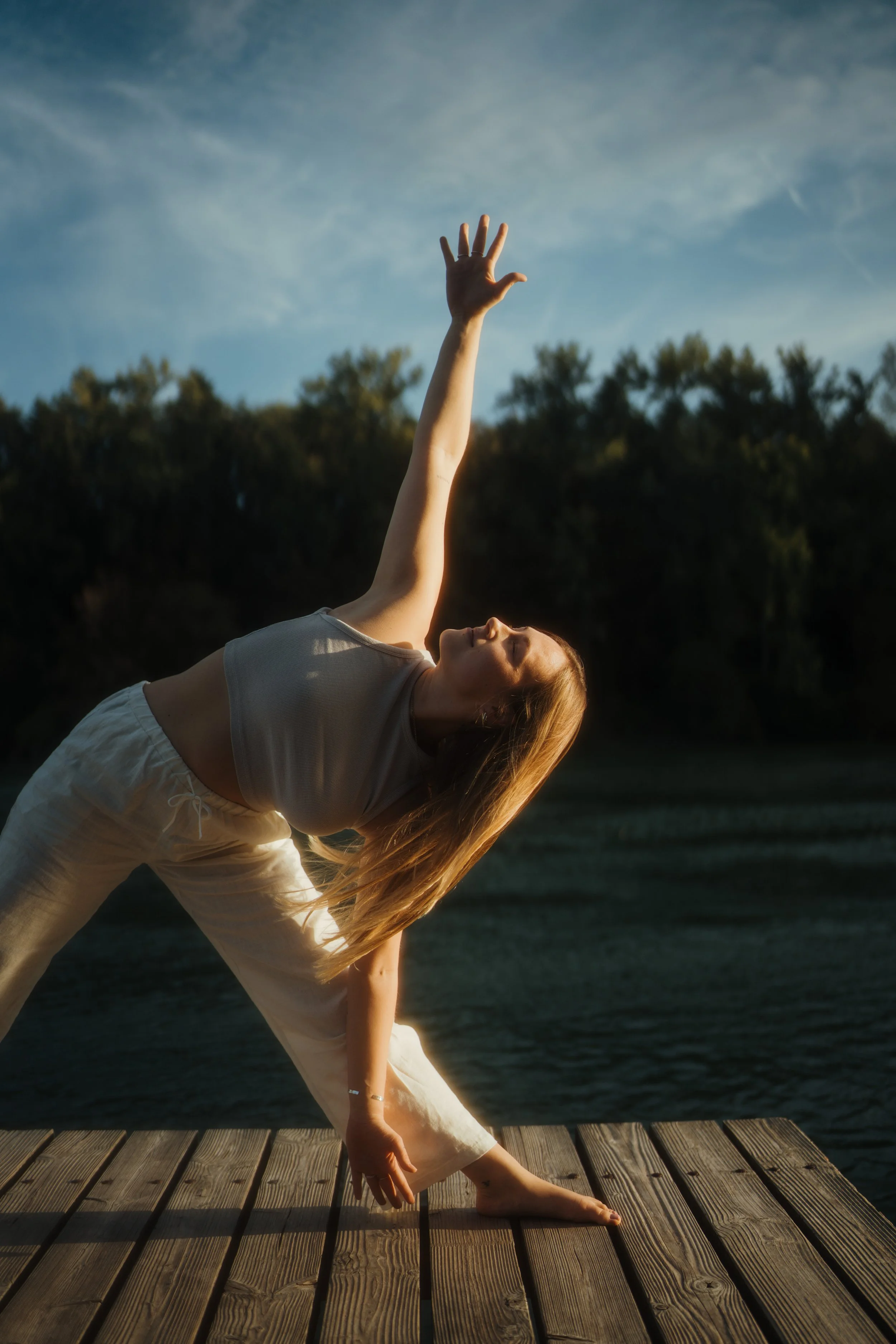 Eine junge Frau macht Yoga auf einer Holzplattform am Wasser, mit Bäumen im Hintergrund und einem blauen Himmel mit einigen Wolken.