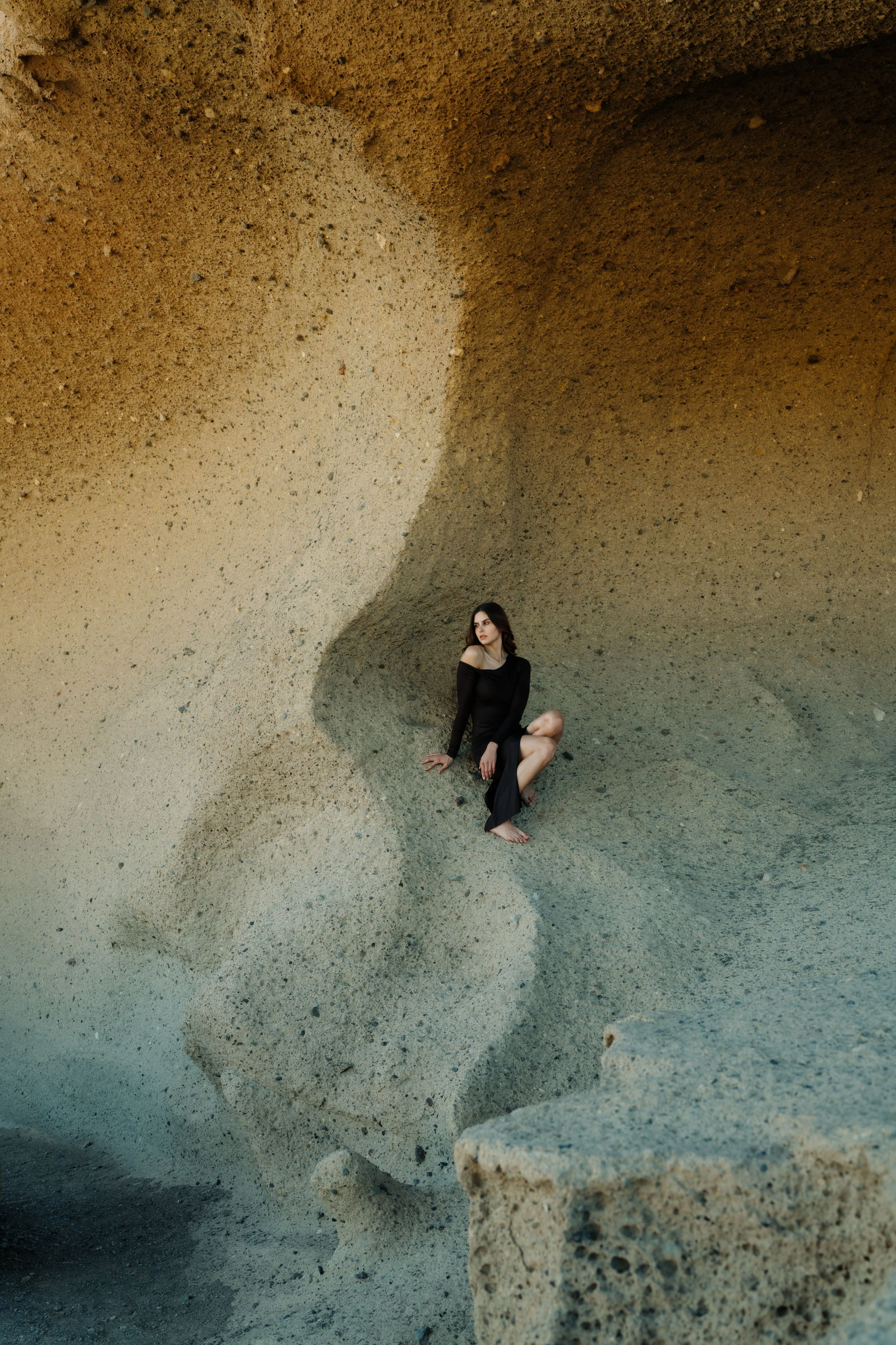 Junge Frau in einem schwarzen Kleid sitzt in einer Felsgrotte mit glatten, sandfarbenen Wänden.