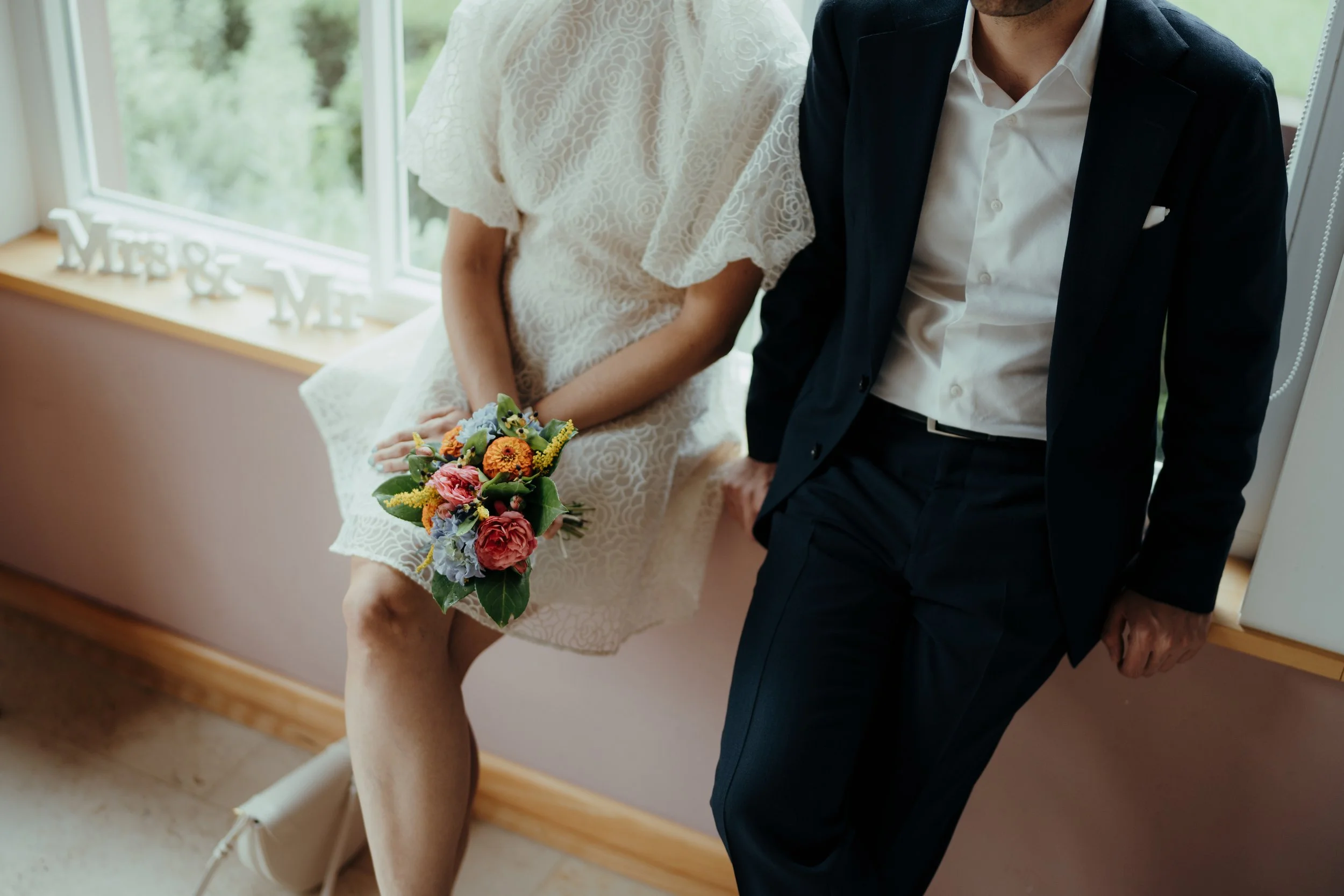 Ein Brautpaar, die Braut trägt ein weißes Spitzenkleid und hält einen Blumenstrauß, während der Bräutigam in einem dunklen Anzug steht, in einem hellen Raum mit großer Fensterfront.