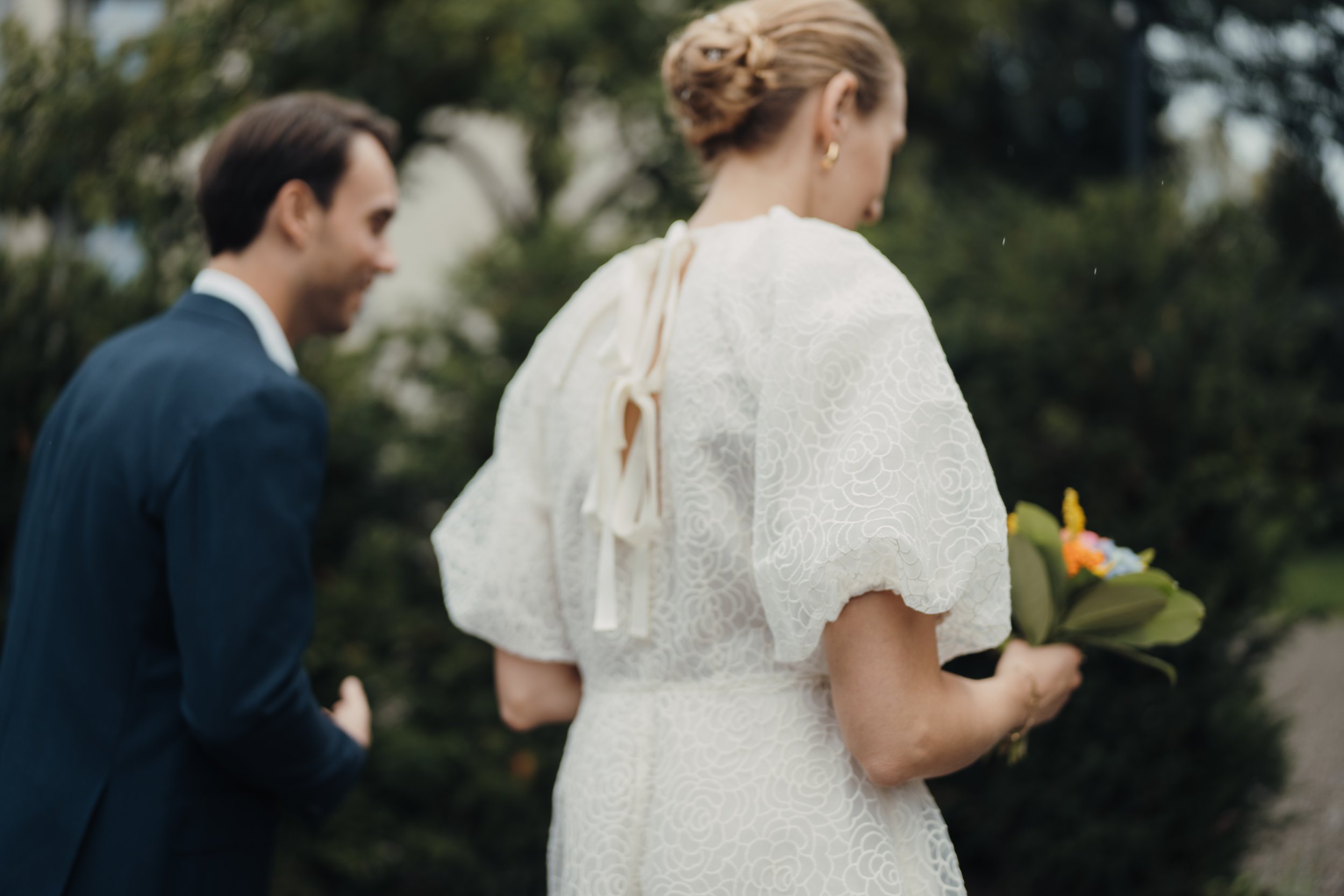Ein Brautpaar bei einer Hochzeit im Freien, die Braut trägt ein weißes Kleid mit Muster und die Umgebung ist grün mit Bäumen.