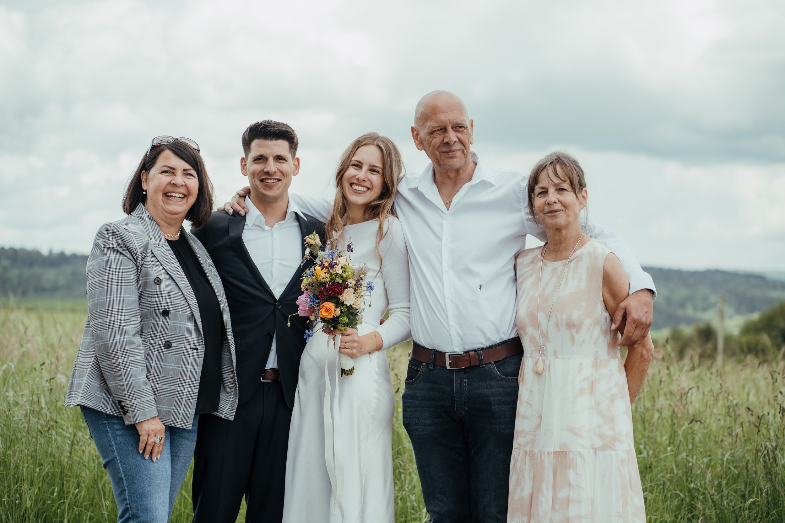 Eine Gruppe von fünf Menschen steht im Freien bei bewölktem Himmel, lacht und umarmt sich, während die Frau in der Mitte ein Blumenbouquet hält.