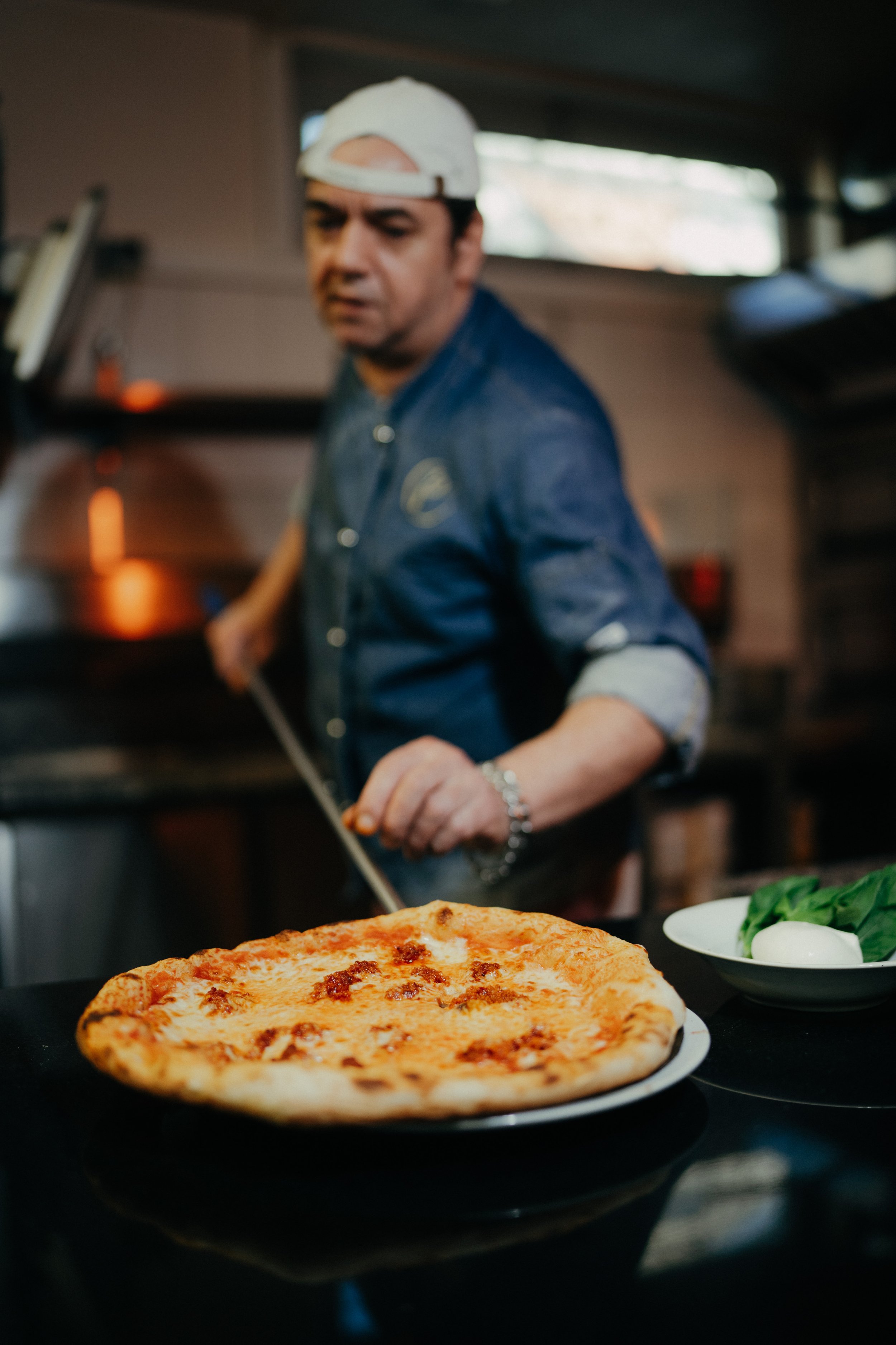 Mann in blauer Jacke und weißem Cap behindert Pizza im Ofen, im Vordergrund eine Pizza auf einem schwarzen Teller, neben einem weißen Teller mit Mozzarella und Basilikum.