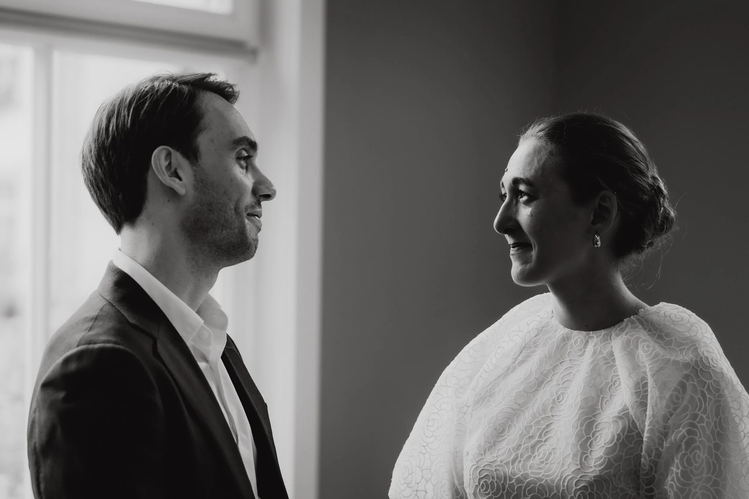 Schwarz-weiß-Foto eines Mannes und einer Frau, die einander gegenüber stehen und lächeln, vermutlich während eines Gesprächs oder eines besonderen Moments, im Hintergrund eine Fensterfront.