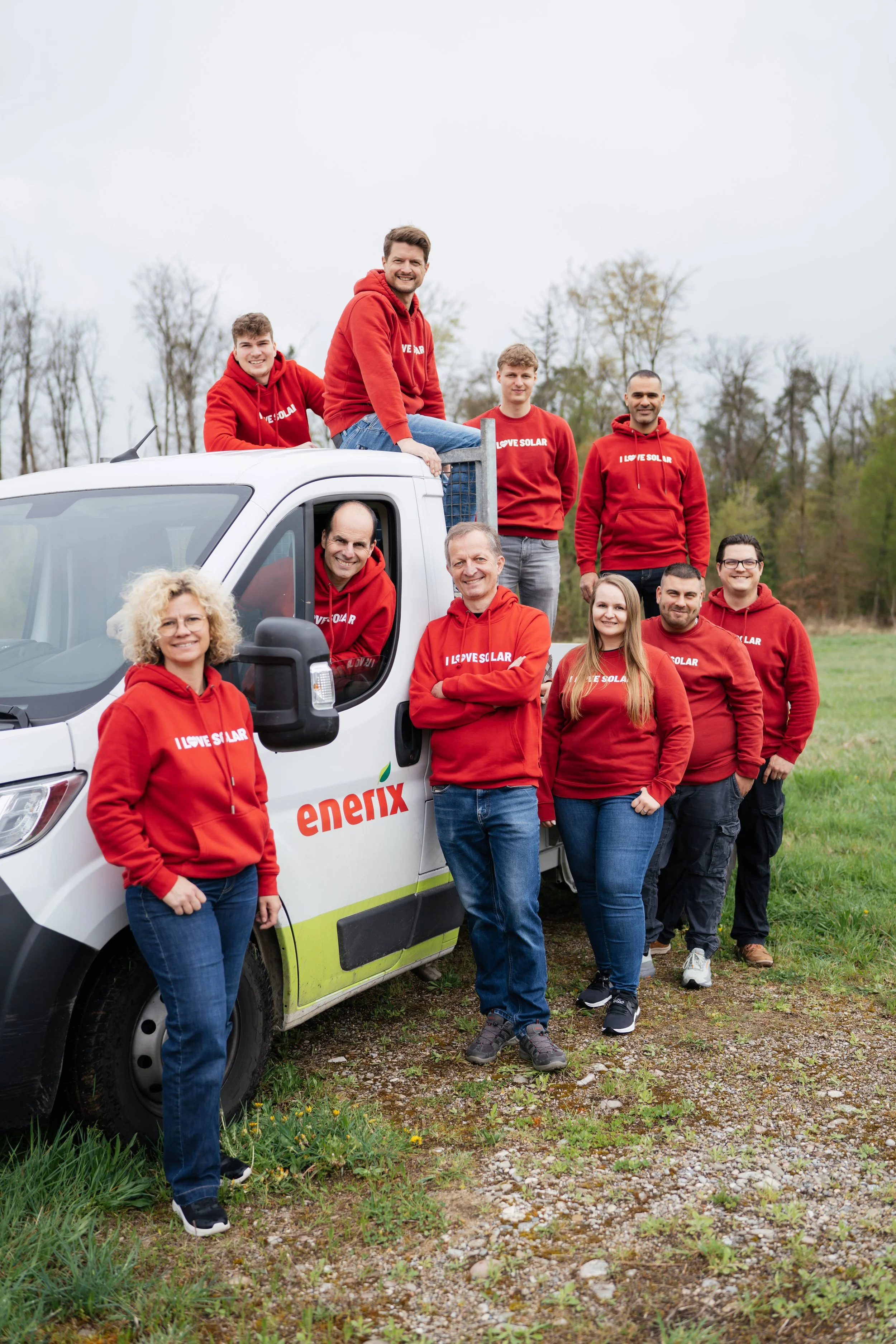 Gruppenfoto von acht Personen, die vor einem weißen Solar-Servicen Fahrzeug stehen, mit einigen auf dem Dach und im und am Fahrzeug. Alle tragen rote Hoodies mit dem Slogan "I LOVE SOLAR". Sie sind im Freien, umgeben von Bäumen und Gras, an einem bew